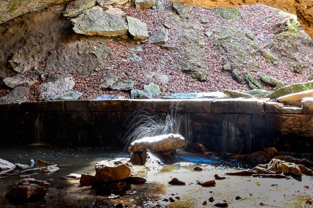 A long-exposure photo inside a rocky grotto. Water drips and splashes off a flat rock and over a low stone ledge into a shallow pool, the moving water blurred into fine silvery streaks and lines. Leaf-covered rocks and a dirt slope rise in the background, with soft light entering from the right.
