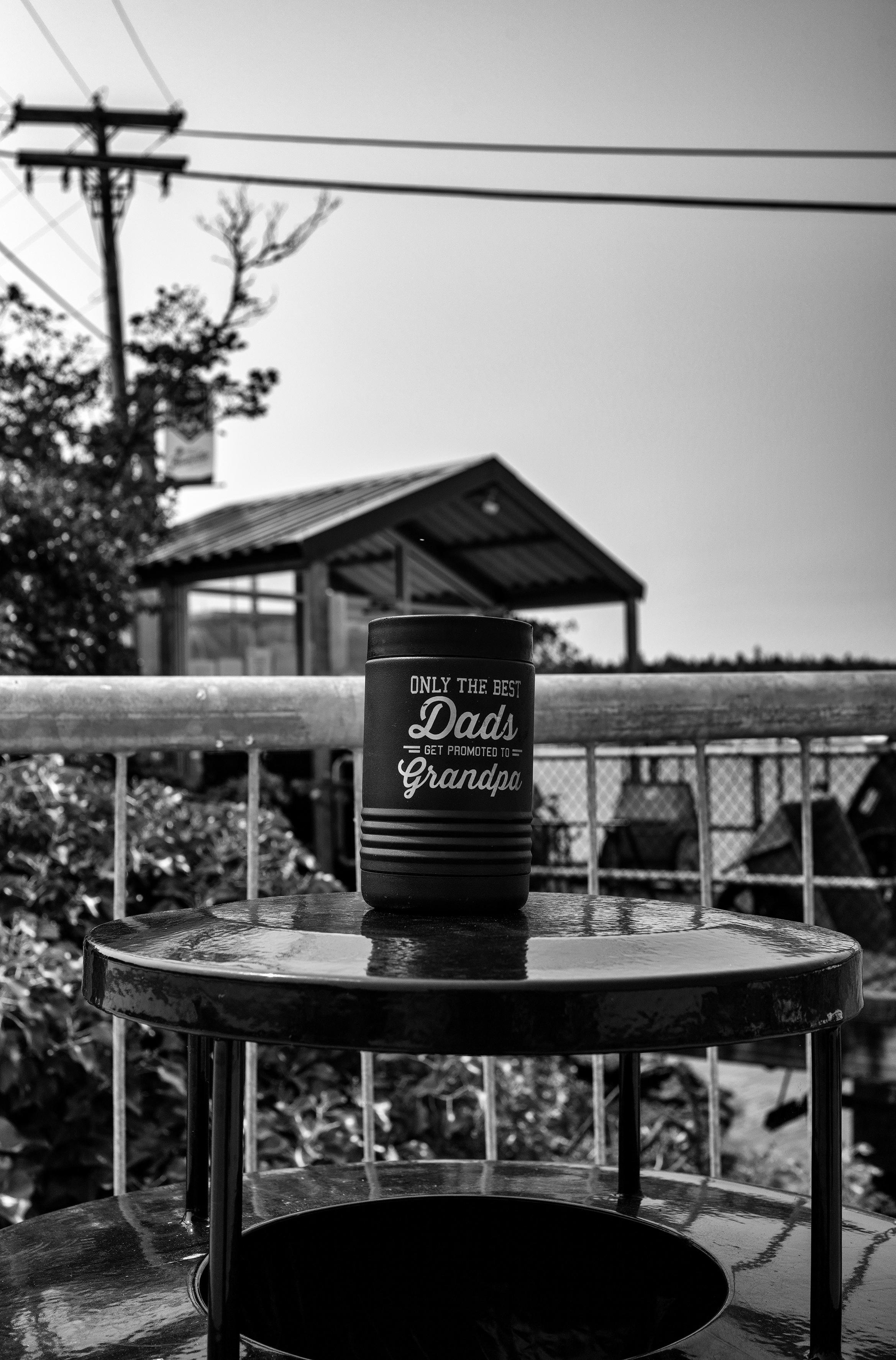 Black and white photo in portrait format of a steel coffee mug forgotten and left behind in a public park on top of a trash receptacle. 

The top part of dark colored cylindrical trash receptacle is centered in the bottom third of the frame. The receptable has a "roof" over the opening to dispose of trash. Sitting on top of the roof is a dark colored handleless steel mug with four ribs at the bottom. "Only the best Dads get promoted to Grandpa" are inscribed above the ribs in white-lettering.

In the background: a steel guardrail running horizontally across the frame directly behind the trash receptacle; a left-tilted power line with power lines running horizontally from left to right across the top of the frame in the far distance; an open gazebo in the middle of the frame in the mid distance; dark colored foliage and trees in starting in the middle of the frame and extending to the top left  in the mid distance.