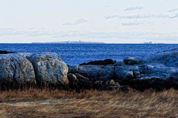 A salt marsh, low granit boulders covered with seaweed and an expanse of ocean separate me from two islands dotted with human built structures visible over the ocean horizon under a drab partially overcast sky