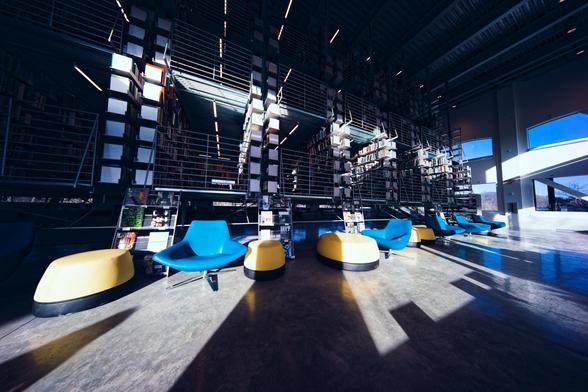 Towards the back are the skewed perspective stacks of a research library suspended from the ceiling over a vast concrete floor with tables and chairs in front of that and dark shadows cast by the edges of windows behind me