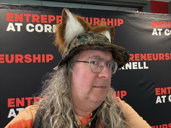 It's me,  looking right at you suspiciusly,  I hope a little canine, i have really long grey curly hair and octagonal glasses and a digital camo boonie cap with a pair of fox ears and behind me a selfie backdrop that reads ENTREPRENEURSHIP AT CORNELL over and over again