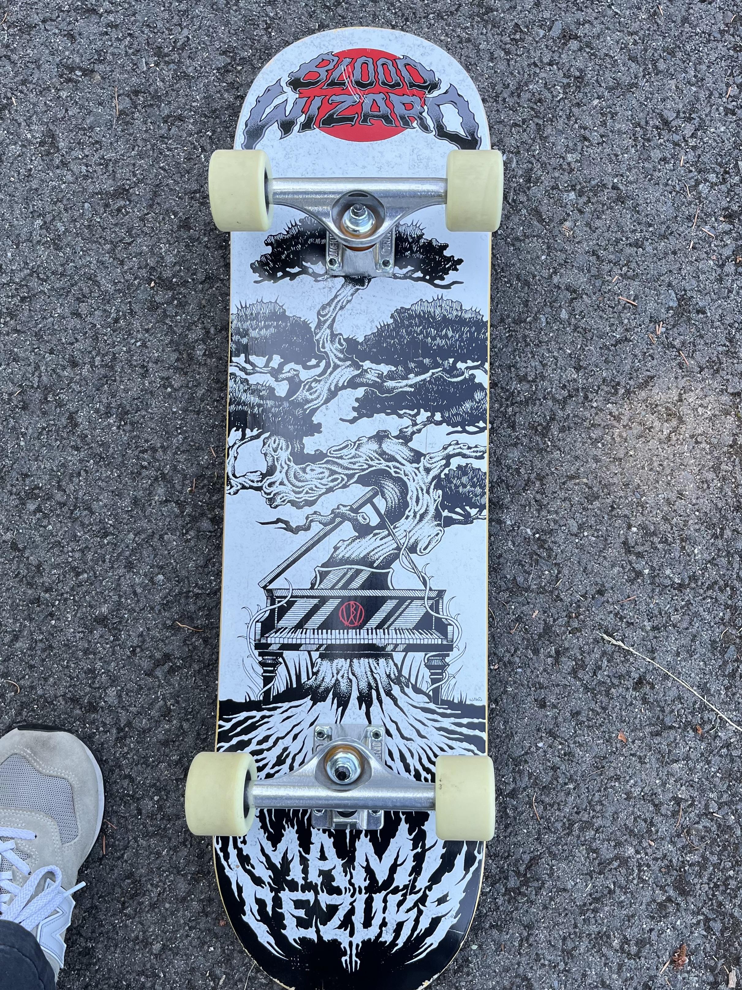 The underside of my Blood Wizard skateboard. It’s a black and white manga-style design of a giant bonsai tree, with a piano sitting under it. On the bottom of the board, a metal font reads “Mami Tezuka”. On the top of the board, similar metal text on top of a red circle reads “blood wizard”.