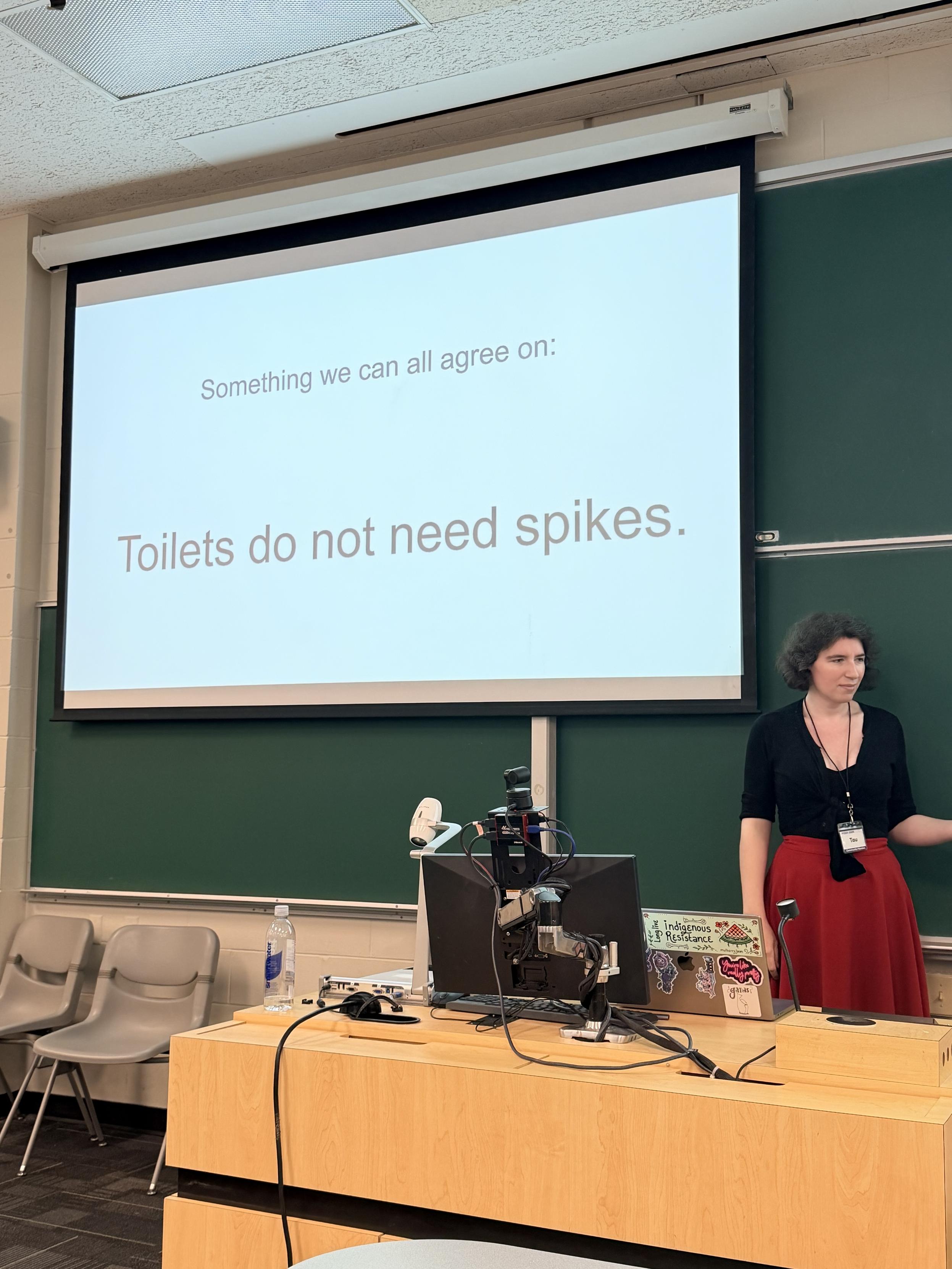 A presentation in a classroom setting, featuring a speaker next to a computer and a projection screen displaying the text: "Something we can all agree on: Toilets do not need spikes." The speaker has curly hair and is wearing a black top.