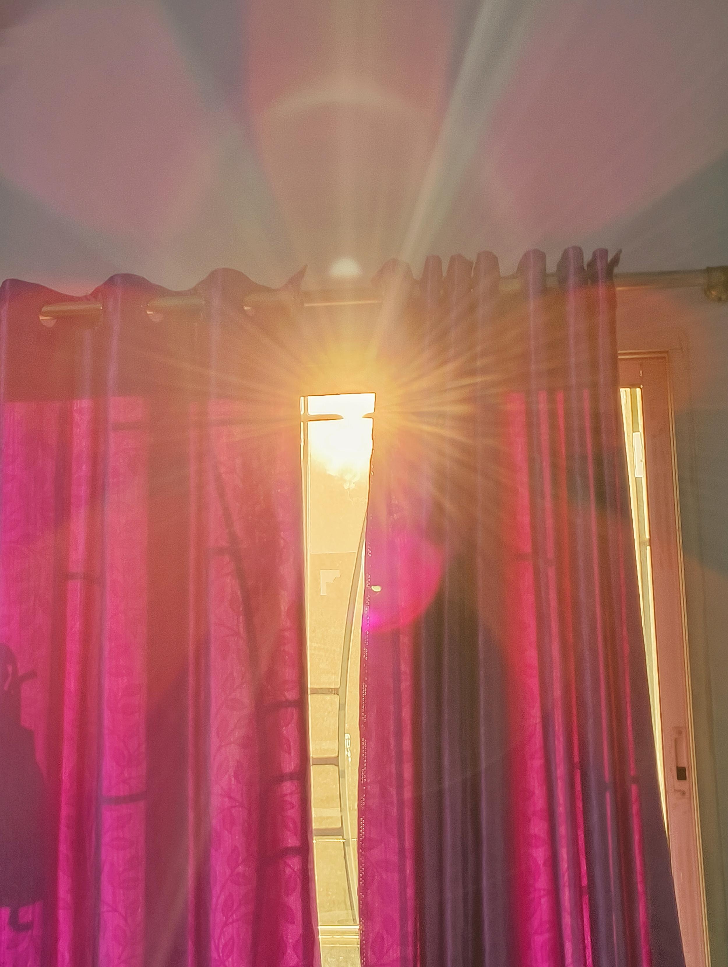 A photograph looking up at vibrant magenta-pink curtains hanging on a silver curtain rod. Bright sunlight pierces directly through a narrow vertical gap between the closed curtains, creating a sharp golden starburst lens flare. Warm rays of light and pink optical flares radiate outward across the ceiling and wall, illuminating the sheer texture of the fabric.