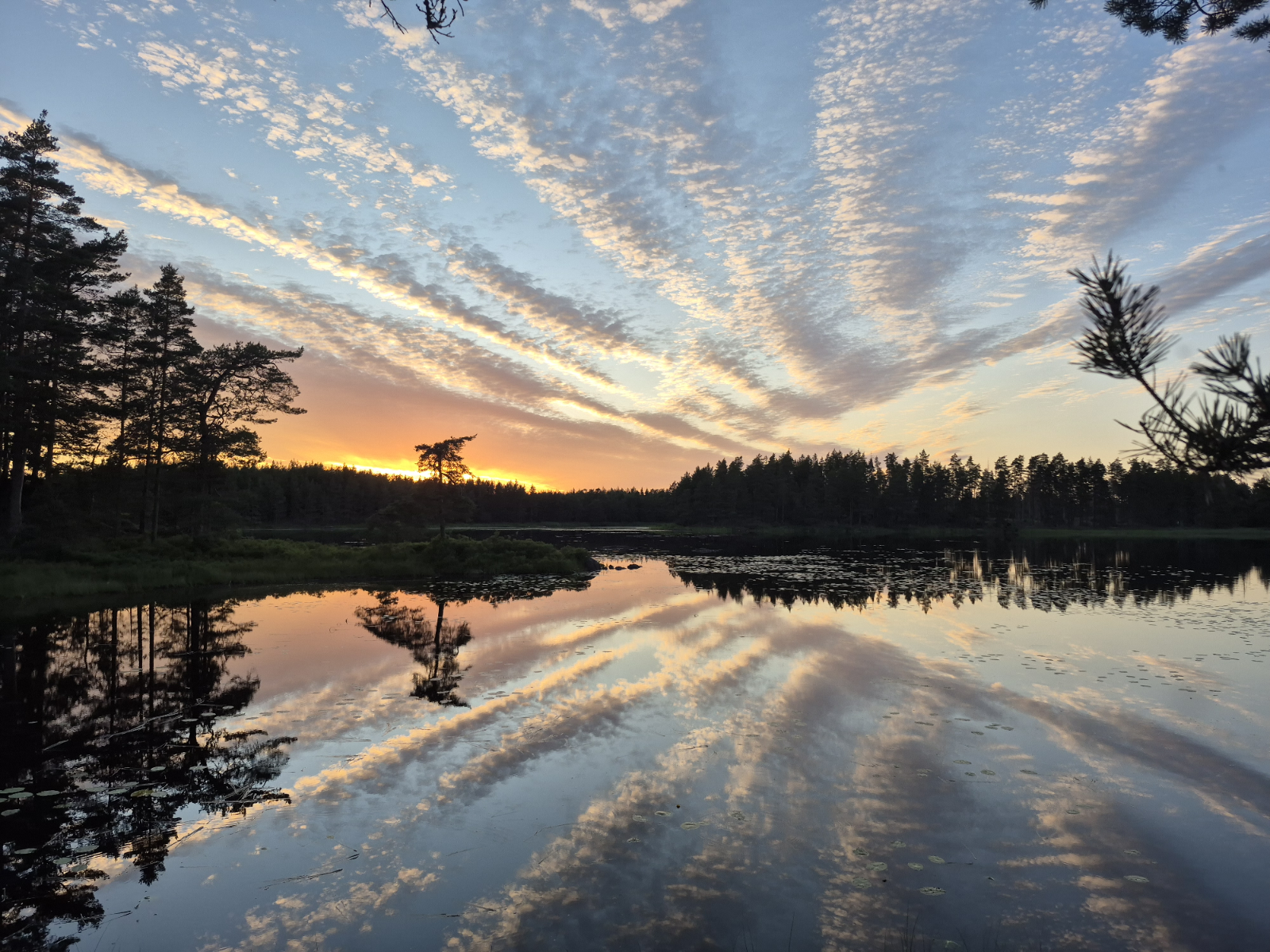 Nach Sonnenuntergang spiegelt sich der Himmel in einem spiegelglatten See. Hinter dem Wald am gegenüberliegenden Ufer strahl das Orange der untergegangenen  Sonne. Über den blauen Himmel erstrecken sich sechs parallel verlaufende Wolkenstreifen. Der Himmel spiegelt sich perfekt in der Wasseroberfläche.