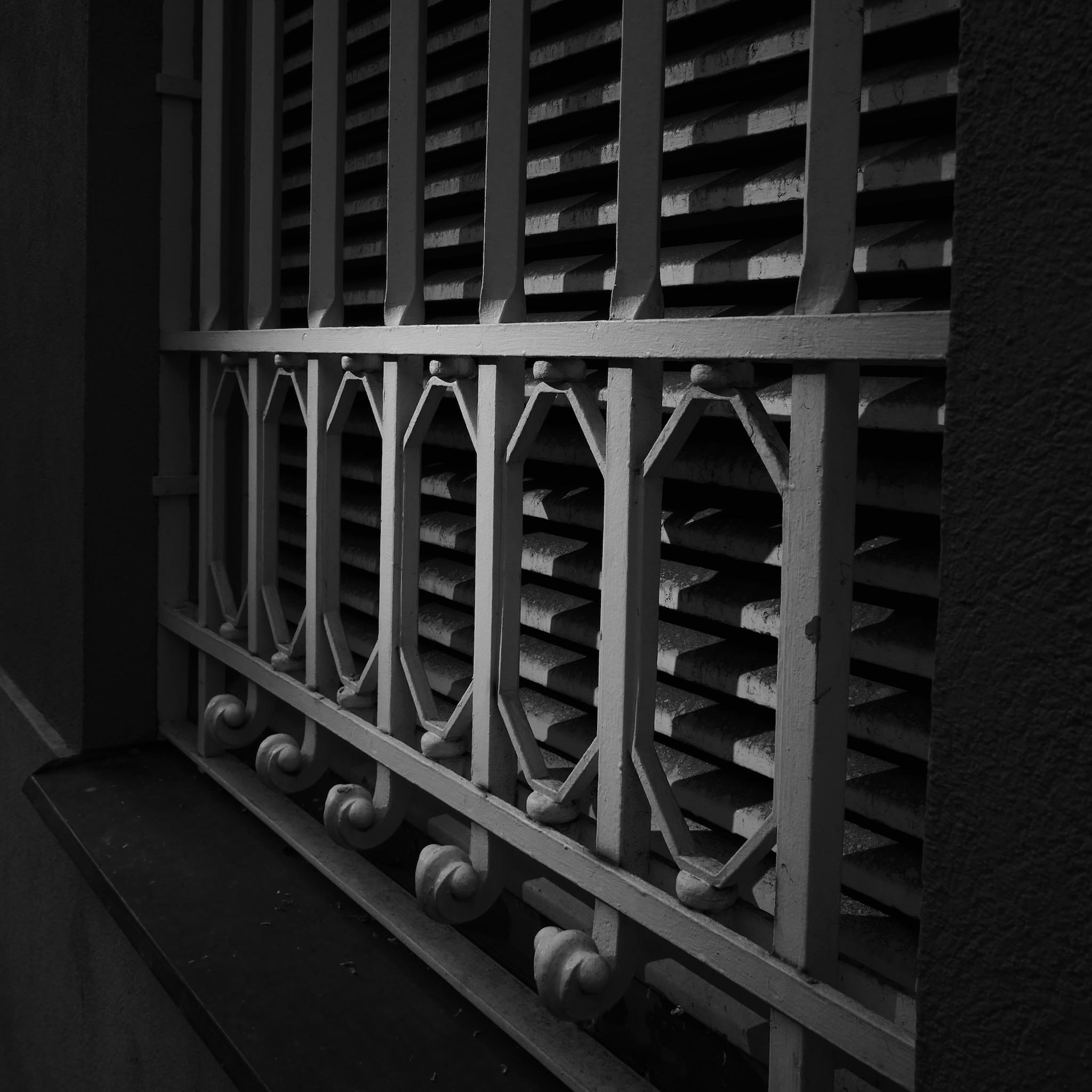 Picture of a closed window. There are blinds behind the glass window. In front of the glass is a metal grid with ornaments. Picture is black and white and has a lot of shadows and contrasts.