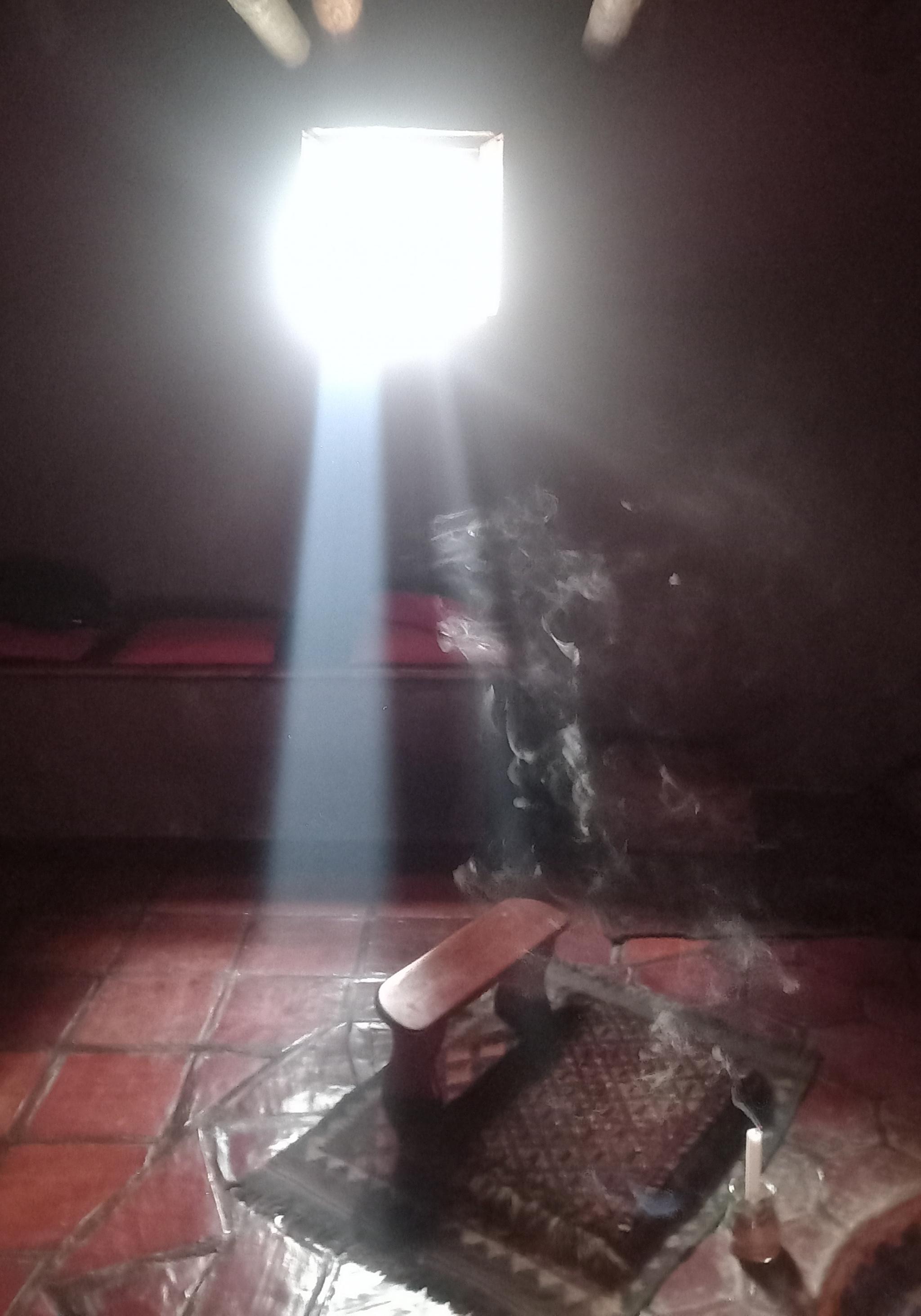 A beam of sunlight illuminates a drifting column of incense smoke 
The floor consists of polished quarry tiles
A kneeling bench is partially lit by the sunbeam.