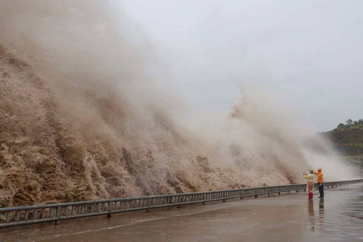 People stand next to huge waves crashing on a waterfront.
