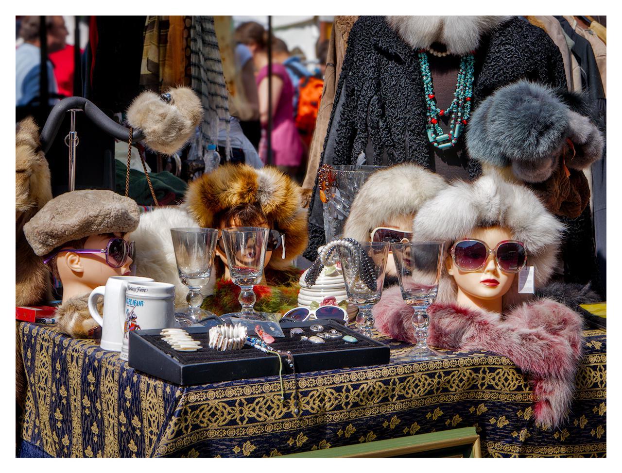 Market stall display featuring several mannequin heads wearing various fur hats and sunglasses, surrounded by glass goblets, jewelry trays, a white mug, and beaded necklaces on a blue and gold patterned tablecloth, with clothing and shoppers visible in the background.