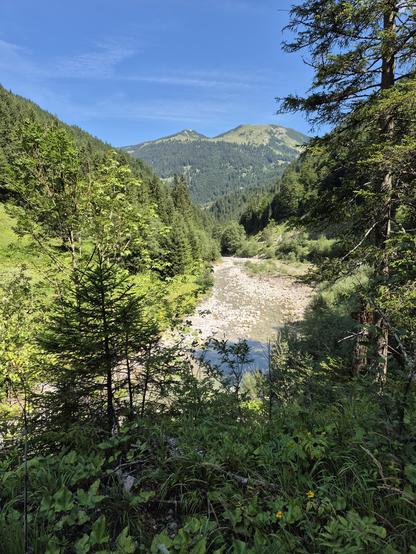This image captures a breathtaking and serene mountain landscape, evoking a sense of tranquility, natural beauty, and adventure. The scene is dominated by a lush, green valley framed by dense forests of evergreen trees. The trees, with their vibrant green foliage, create a rich and textured tapestry that stretches up the slopes of the surrounding mountains.

A **rocky riverbed** cuts through the center of the image, its surface a mix of smooth stones and patches of water, adding a sense of move…
