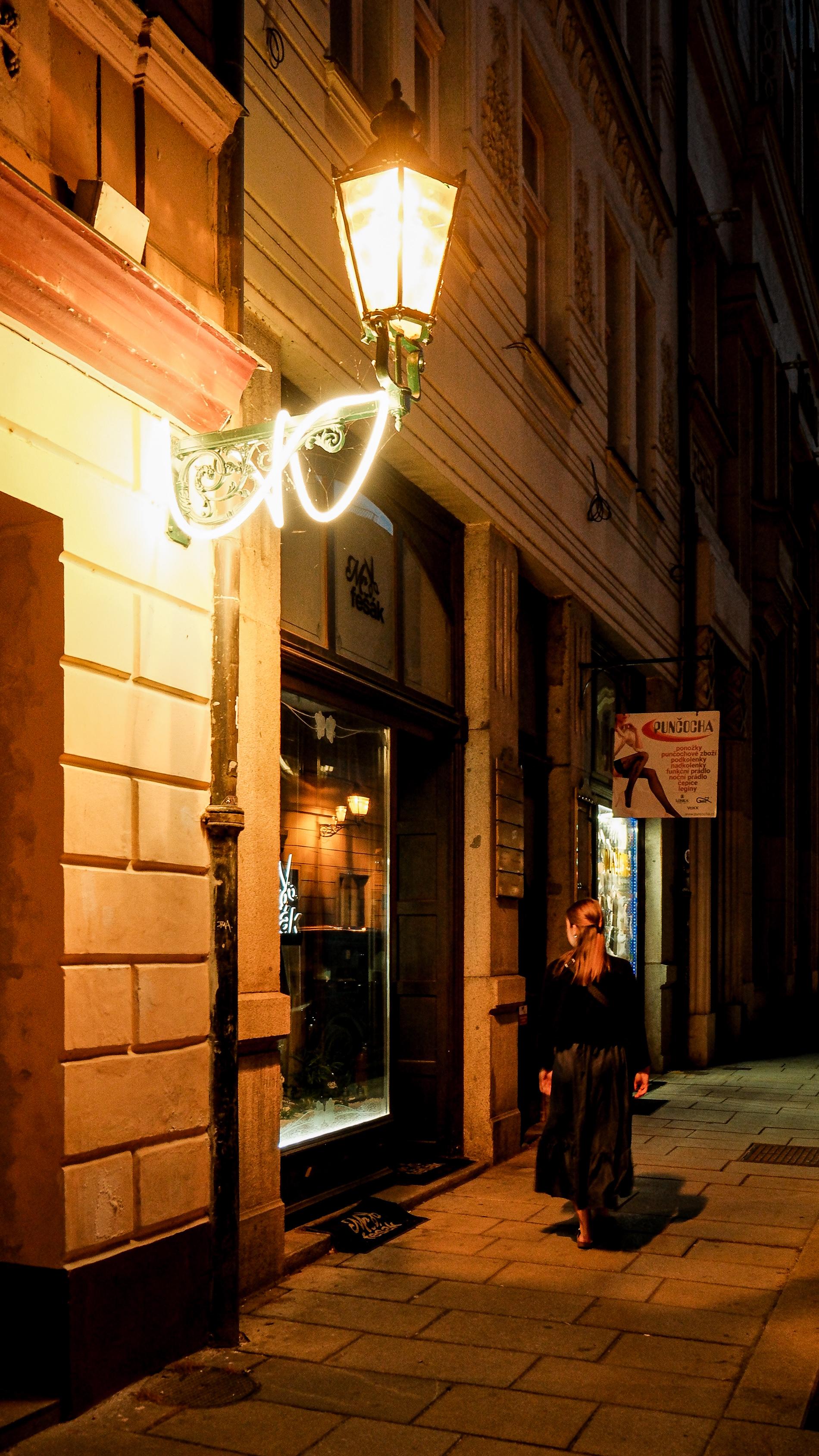 ​A night shot of a narrow cobblestone street. A woman with dark clothes walks past a brightly lit shop window under the warm glow of a vintage wall lantern.