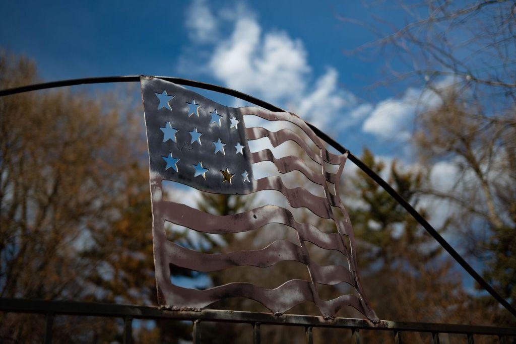A cut-metal American flag sculpture, styled as if waving, with star cutouts in the upper corner that let the sky show through. It is mounted on a black iron archway over a gate. Behind it is a blue sky with soft white clouds and out-of-focus bare and evergreen trees. The metal has weathered, muted tones.