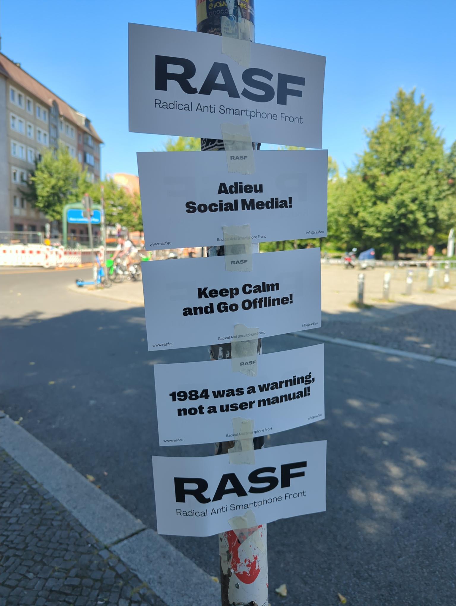 Verschiedene Flyer an einer Straßenlaterne. Auf dem obersten steht RASF, darunter adieu social media, keep calm and go offline, ...