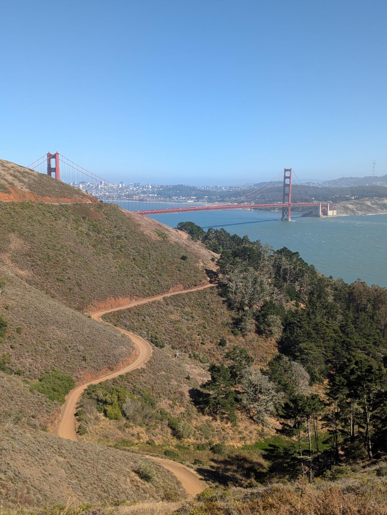 Photo that is probably half blue sky and just under one-quarter deep blue-green water with the remainder being land. In the foreground to the lower left, the land is a steep hill of low green growth with a flat rough cut fire road winding around and tall green trees near the bottom. The water is spanned by a long orange-red suspension bridge. One tower in the distance is completely visible, and the other is partly obscured by the hillside in the foreground. Across the water are several low green hills to the right and the dense outline of a city to the leftm