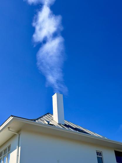 The top of a house with a chimney under a blue sky. It looks like the wispy clouds are coming from the chimney