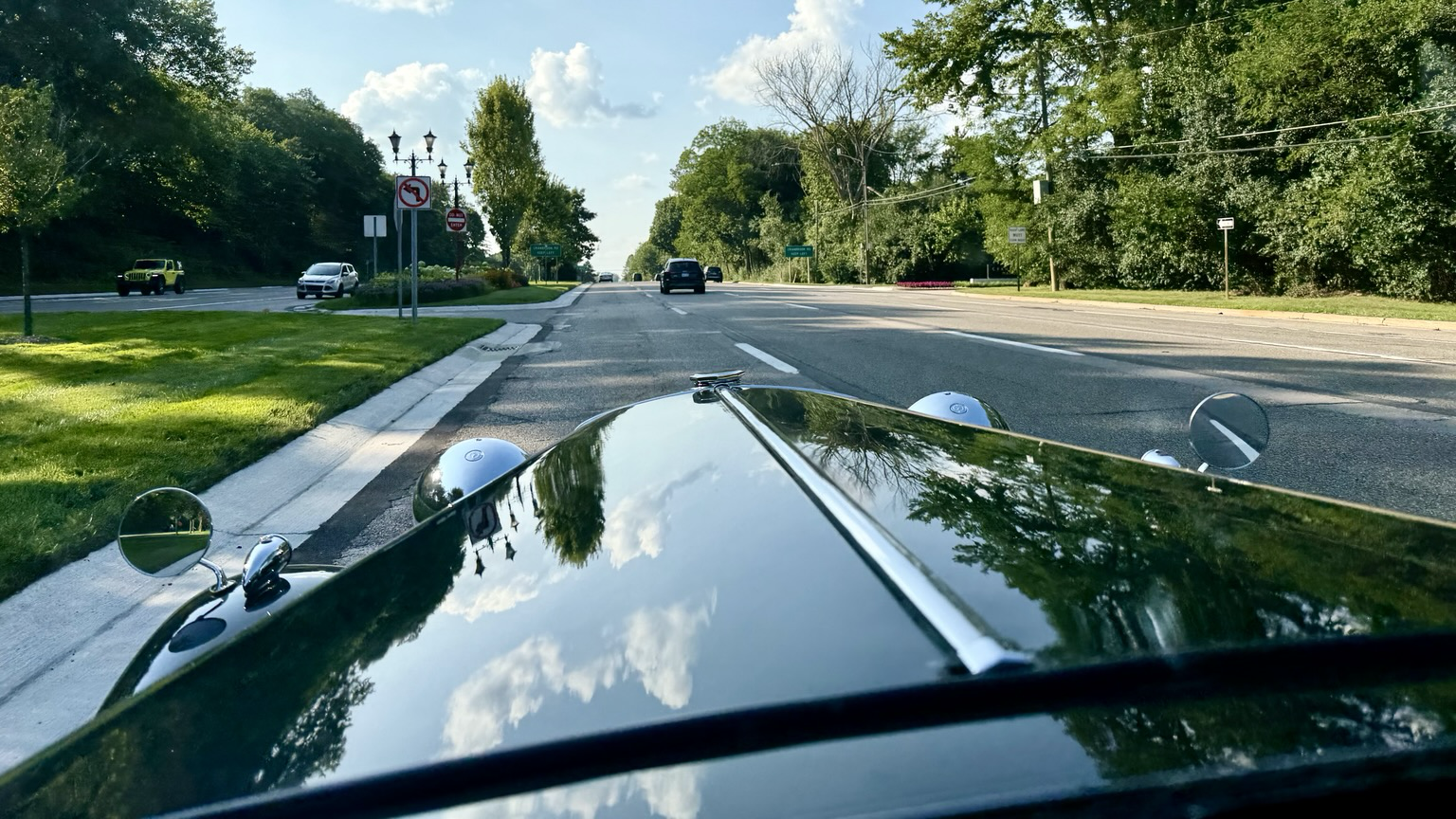 Photograph down the hood of a dark green 1950 MGTD as it drives down a wooded boulevard in the summertime.