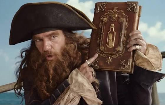 A man dressed as a pirate with a large hat and an elaborate beard is holding a decorative book, pointing at it with a serious expression. The background features a bright sky and visible ocean.
