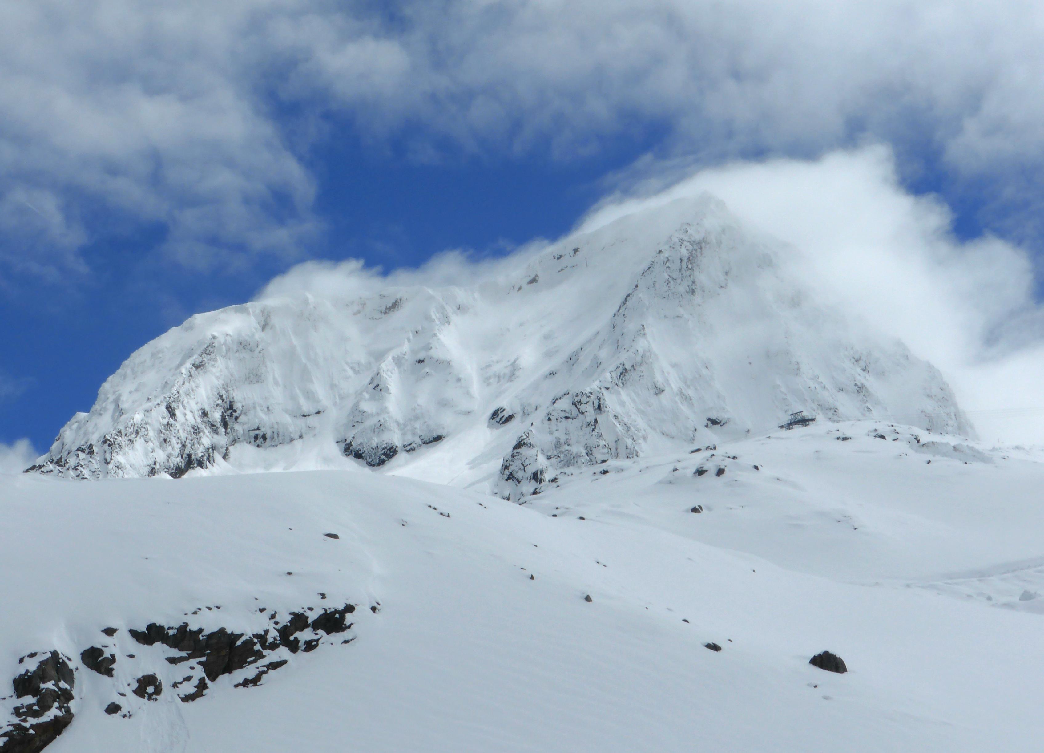 Das Bild zeigt den von Wolken und Schneewehehen umhüllten Gipfel des Zuckerhütl im Stubaital. Hinter der Bergsilouhette bricht der blaue Himmel durch. Stubaital 2015