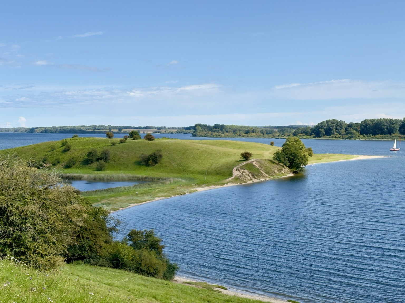 Landschaft mit sanften, mit Gras, Büschen und kleinen Bäumen bewachsen, durchzogen von einem Fluss, der im Vordergrund eine Bucht bildet; alles unter einem blauen Himmel mit wenigen Wolken am Horizont