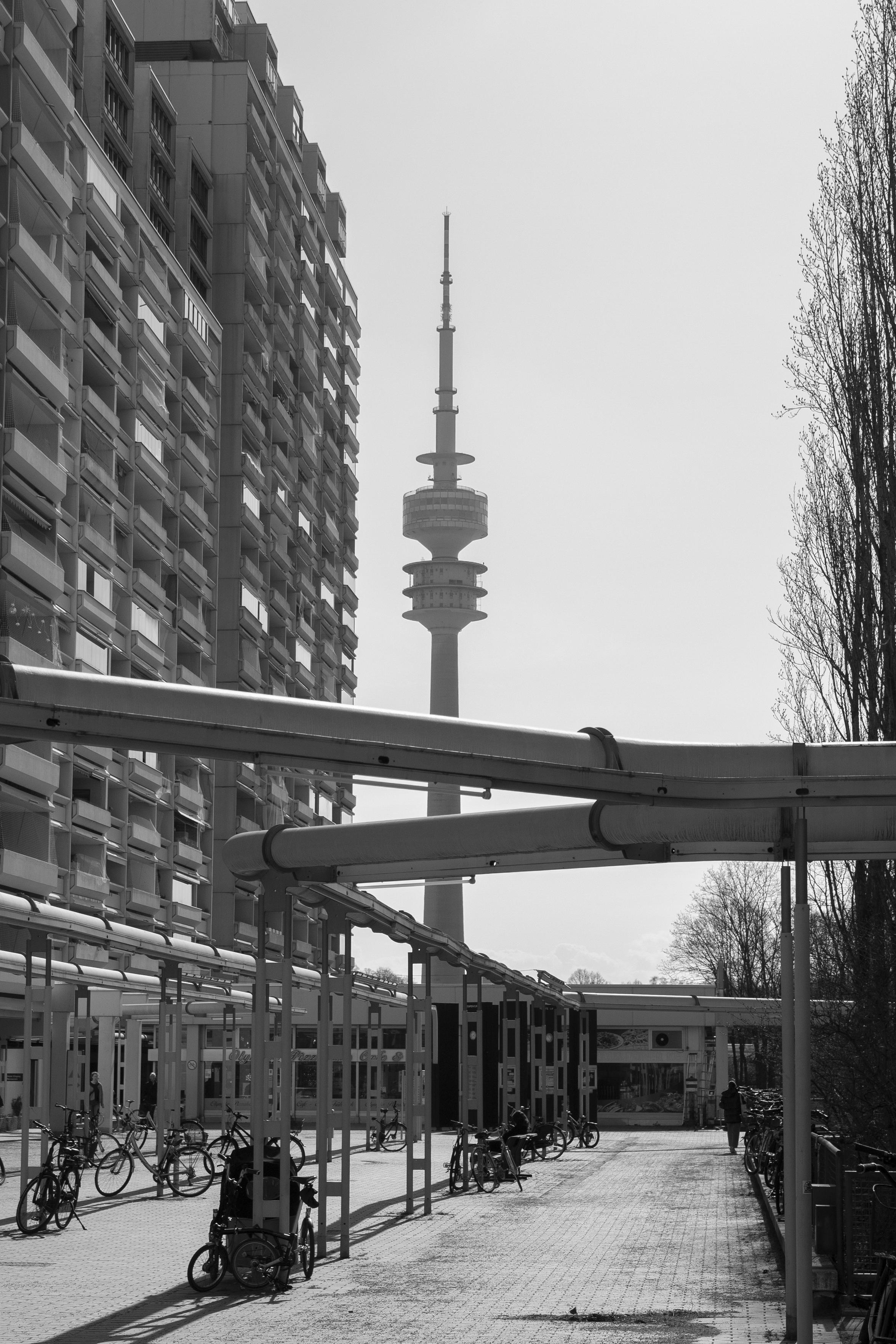 The Munich Olympiaturm, an 291,28m tall television tower is in the center of the photo. The camera points at it from a few hundred meters distance at ground level. To the left of the tower there's a tall multistory residential concrete building building with many balconies slightly angled to the line of sight, appearing as tall as the tower because it's so near. Above the paved ground there's elevated thick metal piping pointing towards the tower, possibly gas or water pipes. The pipe supports are surrounded by parked bicycles.