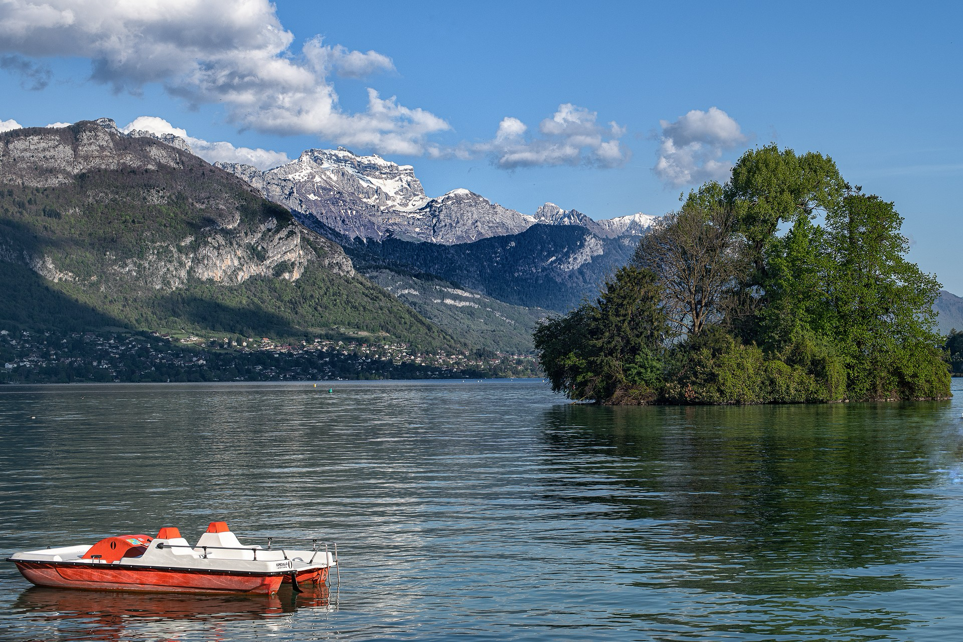 Bildbeschreibung (Deutsch) Das Bild zeigt eine friedliche und malerische See- und Berglandschaft:  * Das Hauptmotiv: Im linken Vordergrund schwimmt ein kleines, leeres Tretboot in den Farben Weiß und leuchtendem Orange sanft auf dem ruhigen Wasser.  * Die Umgebung: Das Boot treibt auf einem weitläufigen See. Im rechten Mittelgrund ragt eine kleine, dicht mit grünen Bäumen bewachsene Insel aus dem Wasser. Der Hintergrund wird von einer majestätischen Bergkette dominiert, deren höchste Gipfel noch mit Schnee bedeckt sind. Am Ufer am Fuße der Berge erstreckt sich eine weitläufige Ortschaft unter einem strahlend blauen Himmel mit weißen Schönwetterwolken.  * Die Stimmung: Die Szene strahlt eine unglaubliche Ruhe und eine idyllische Urlaubsatmosphäre aus. Das harmonische Zusammenspiel von friedlichem Wasser, üppiger Natur und der imposanten Bergkulisse lädt zum Durchatmen und Entspannen ein. Image Description (English) The picture shows a peaceful and picturesque lake and mountain landscape