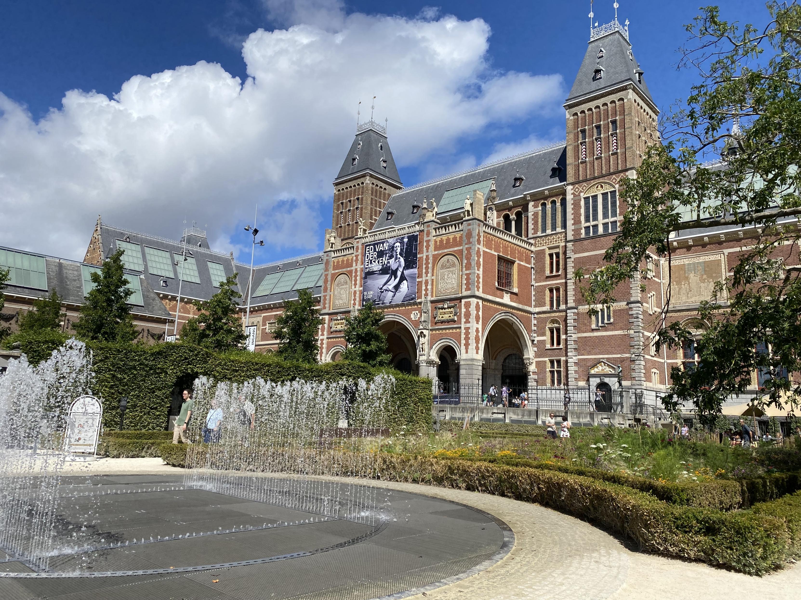Blik op het Rijksmuseum vanuit de tuin. Op de voorgrond een fontein. Lucht is blauw, met wolkje.