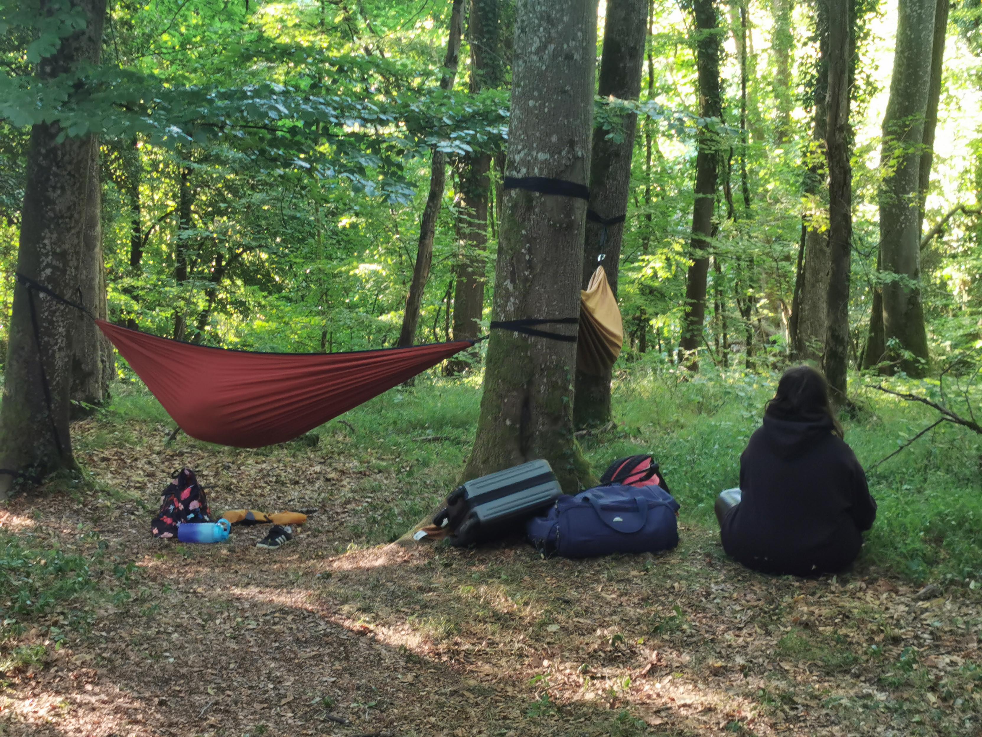 En forêt, un hamac est installé et une jeune fille assise de dos ouvre un sac.