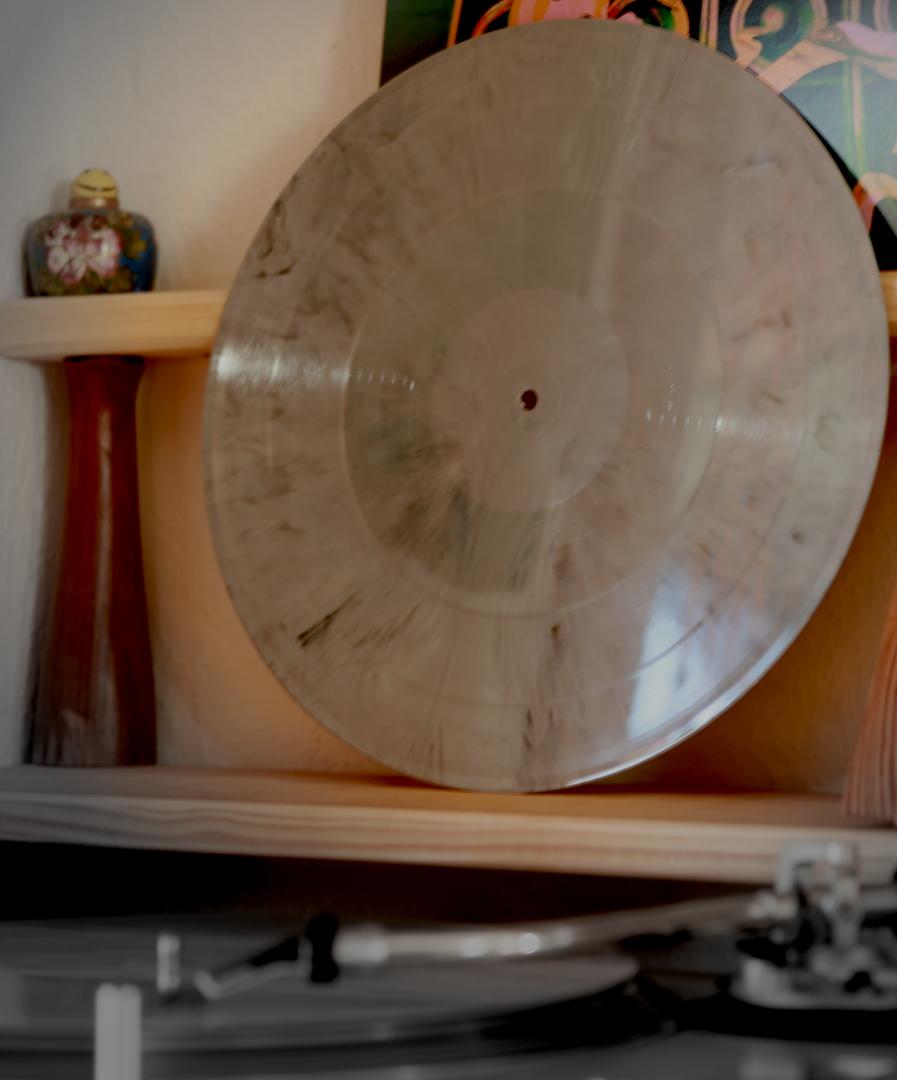 The vinyl records from the album "Soundboy’s Gravestone Gets Desecrated By Vandals", one standing and the other lying on a turntable. They are grey marbled and have no label. One is standing on a wooden base behind and above the turntable, the other is lying on the turntable at the bottom of the picture, the lower part of the image is out of focus. A record sleeve can be glimpsed behind the top vinyl record. In the top left-hand corner, on a wooden board, stands a small Italian vase containing a grinning smiley ball.