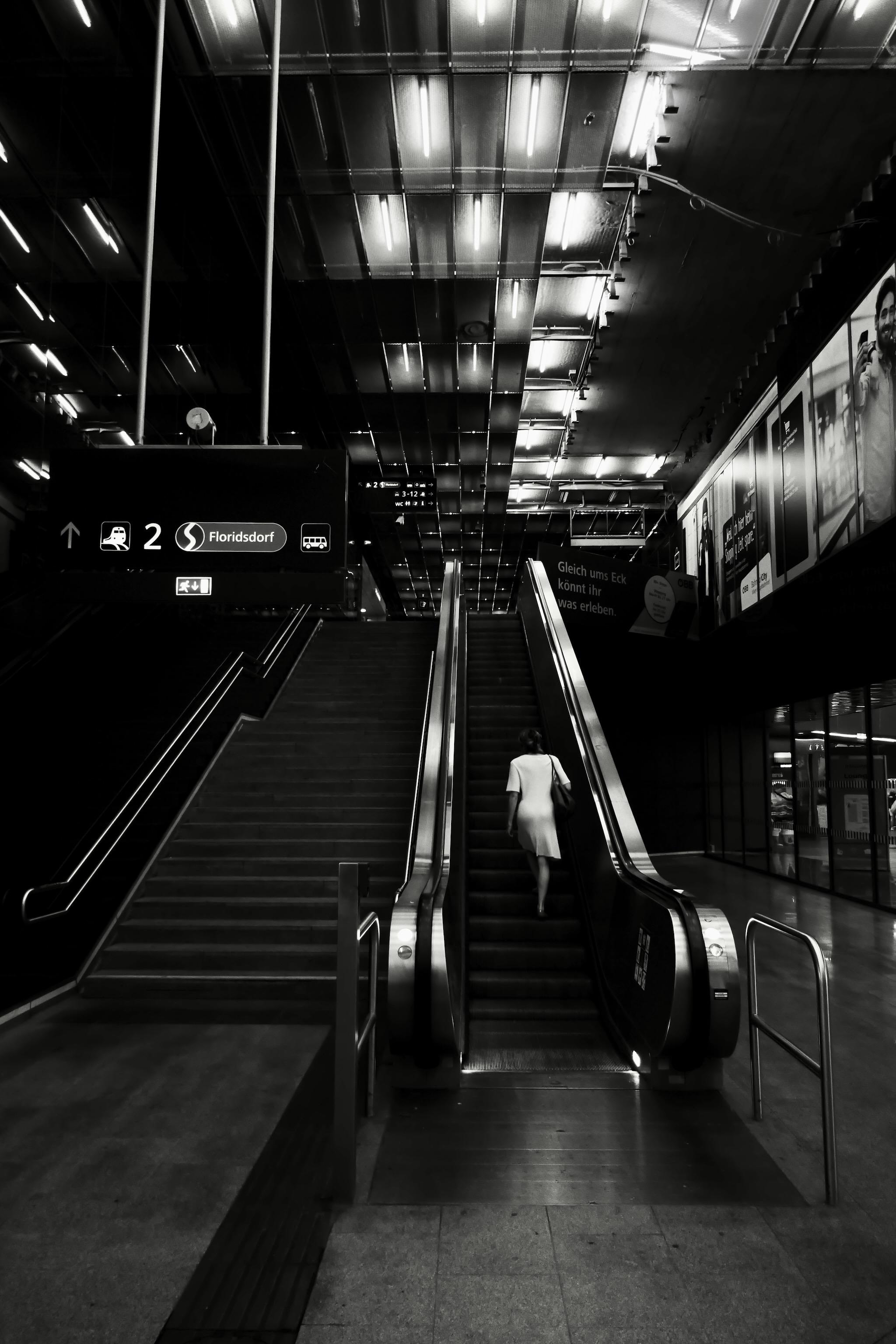 Photo shows an escalator  in a train station going up. A woman is standing on it, alone. White neon stripes are on the roof far up there. On the left side of the escalator is a normal stair. On the right side is an elevator. Picture is black and white.