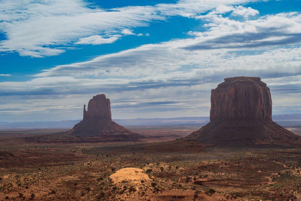 A wide view of Monument Valley under a large sky streaked with white clouds. Two red sandstone buttes stand on the desert plain: East Mitten Butte on the left, with a slender thumb-like spire, and the broad, fortress-like Merrick Butte on the right. Red desert scrub and a pale sand dune fill the foreground, with hazy mesas on the distant horizon.