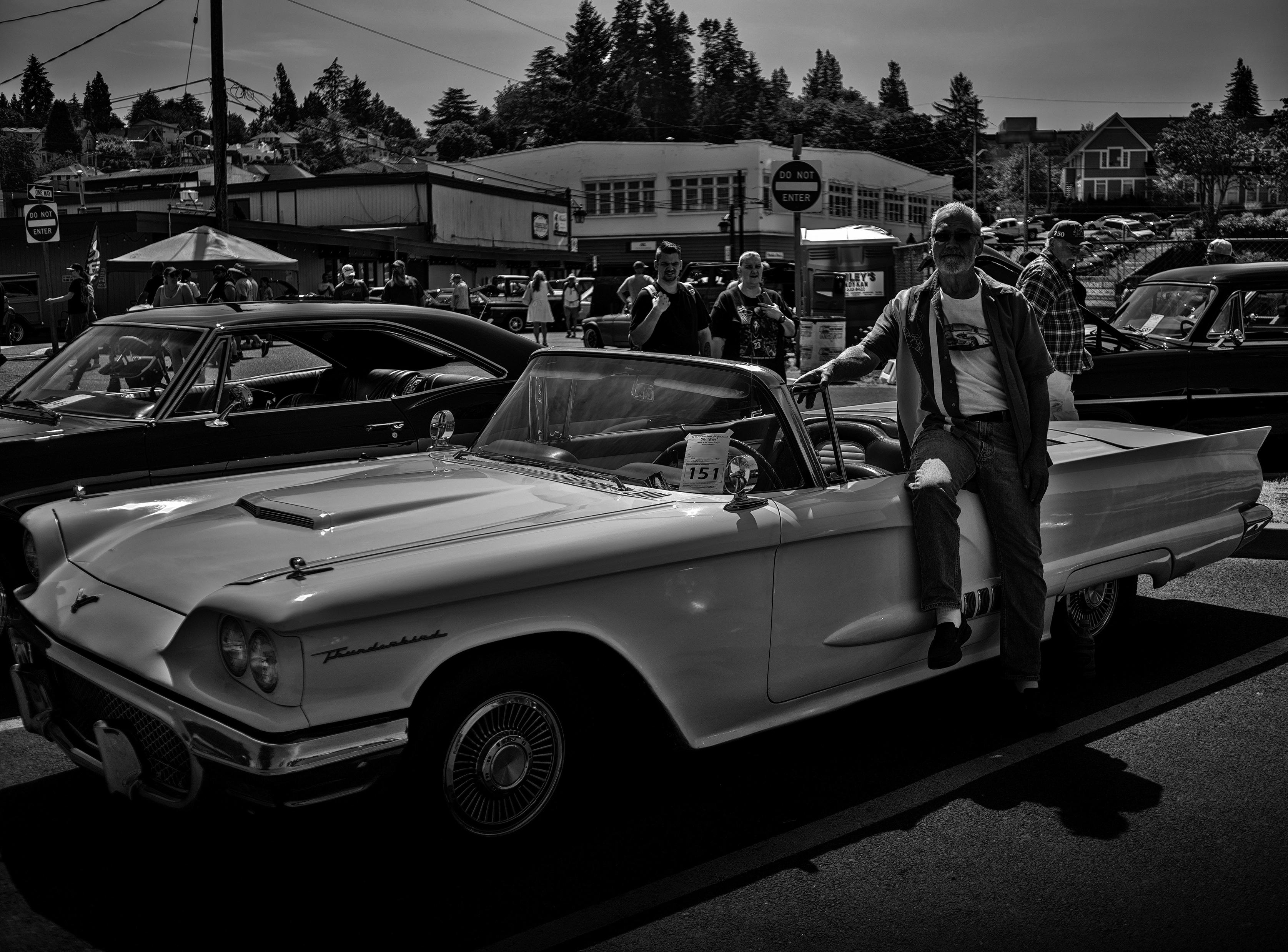 Black and white photo in landscape format of the proud owner of a white convertible 1958 Ford Thunderbird.

John is dressed in dark steel framed rectangular sunglasses, white T-shirt with a Ford Thunderbird printed across the chest, dark colored unbuttoned collared shirt over the T-shirt denim jeans, and black shoes.

Before taking the photo John asked to give him a couple minutes to close the trunk and engine, and retract the convertible's soft-top roof. He then propped himself up on the driver side door with right hand placed on the small side window, right knee bent over the door, and left left planted on the ground for balance.

Groups of spectators to the car show, a number of other cars on display, and a typical US small town business and residential structures are in the background.