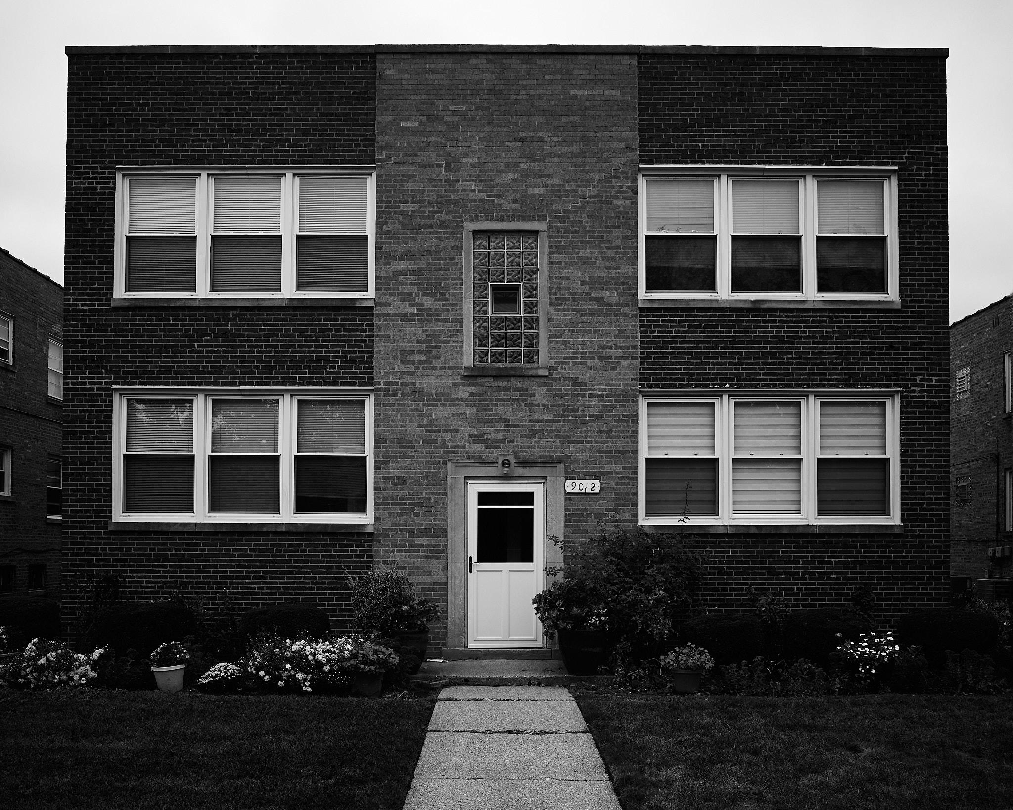 A two-story brick apartment building is centered in the image. It features multiple windows, some with blinds, and a white door at the front. A concrete pathway leads to the door, flanked by flower pots and low shrubs in the foreground.