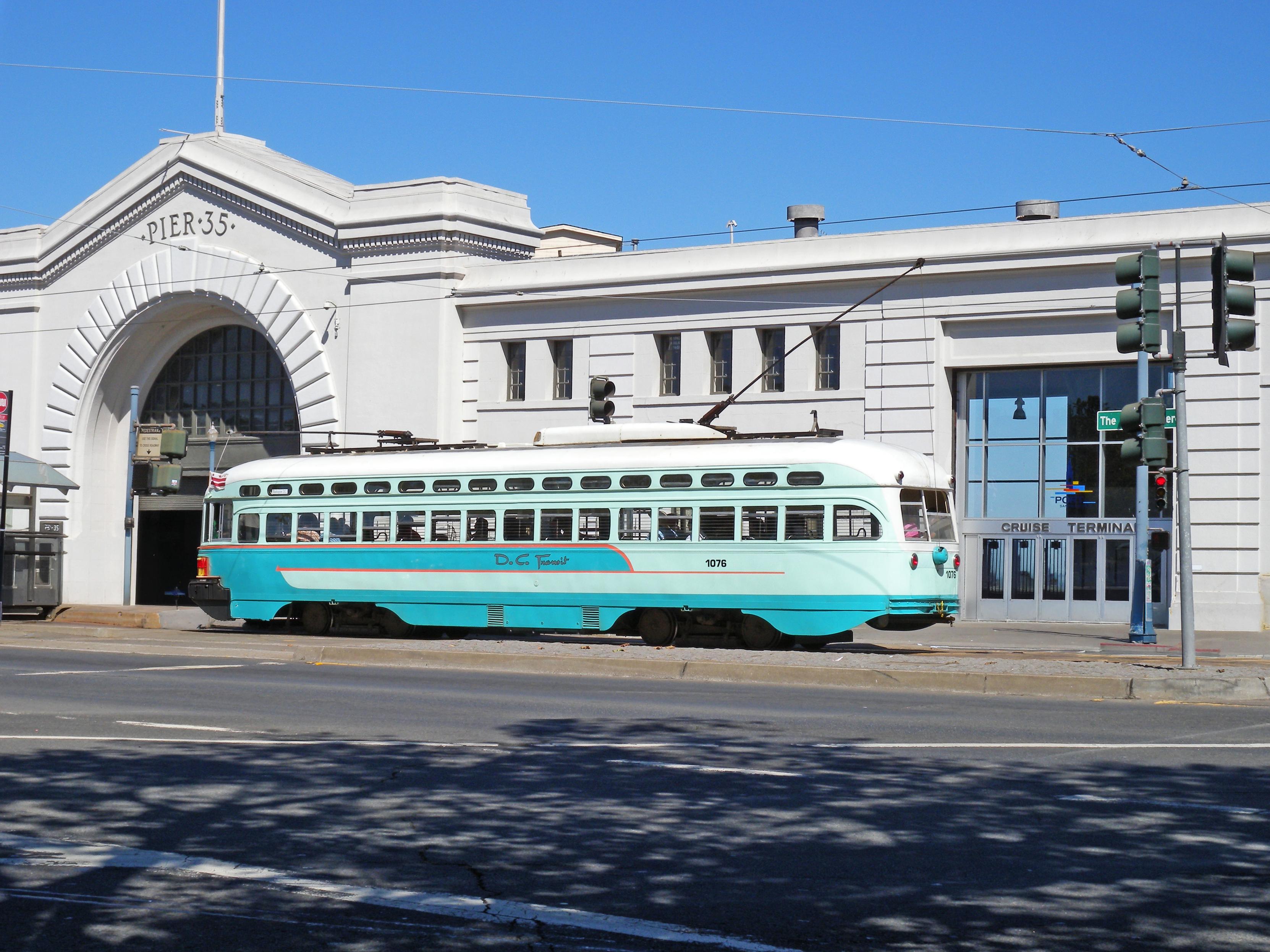 This is a historic PCC streetcar from the San Francisco Municipal Railway, model number 1076, shown here in its Washington, D.C., livery. The streetcar runs on the F Line (Market & Wharves) along the Embarcadero. The background shows the Pier 35 Cruise Terminal in San Francisco.