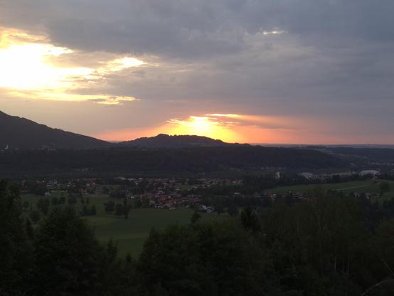 The image captures a serene sunset over a hilly landscape. The sky is painted with soft hues of orange, pink, and purple, as the sun dips behind a distant mountain, casting a warm glow. The foreground features lush green fields and scattered trees, leading to a small village nestled in the valley. The silhouette of the hills creates a layered effect, adding depth to the scene. The horizon stretches wide, with faint signs of civilization—buildings and roads—dotting the landscape. The overall moo…