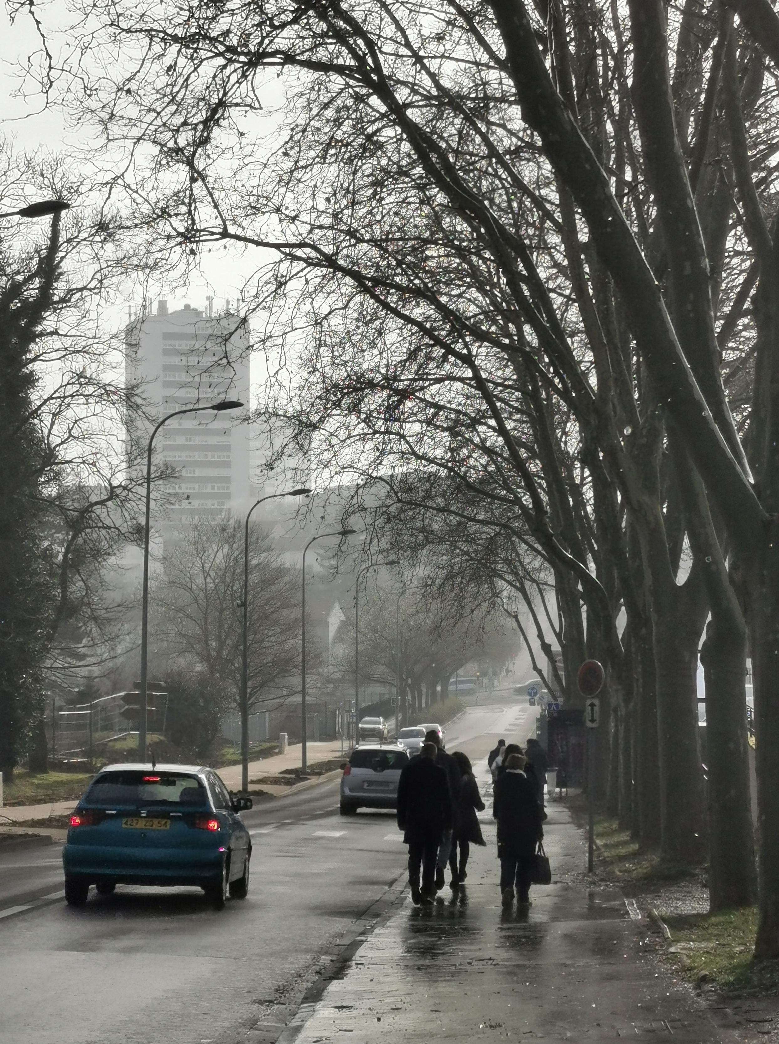 Dans une ambiance de brume lumineuse, un groupe de personnes descend une avenue. Au fond apparaissent fantomatiques des immeubles.