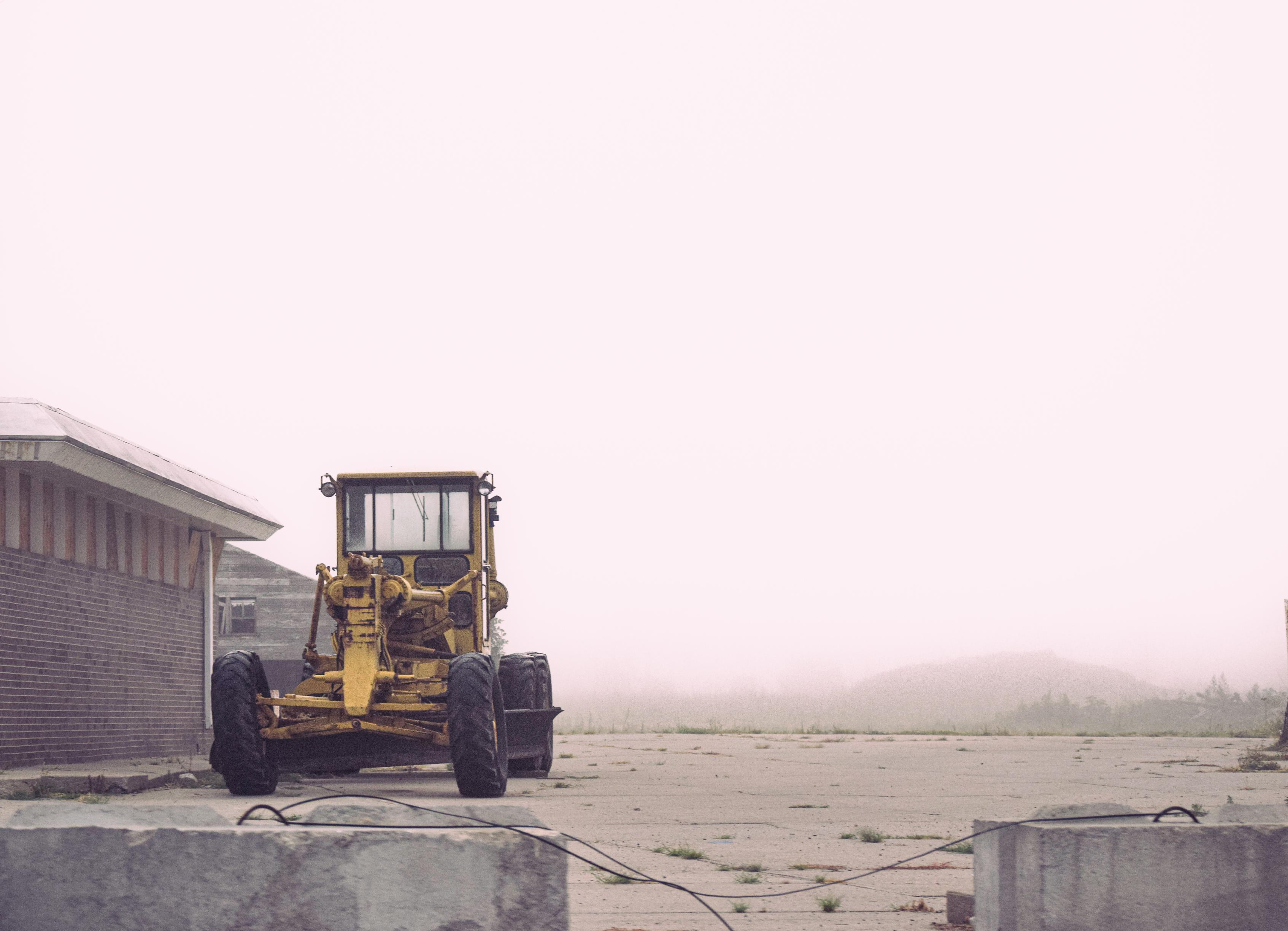 A solitary yellow bulldozer is parked on an overcast day beside a brick building, with misty surroundings creating a sense of isolation and calm.