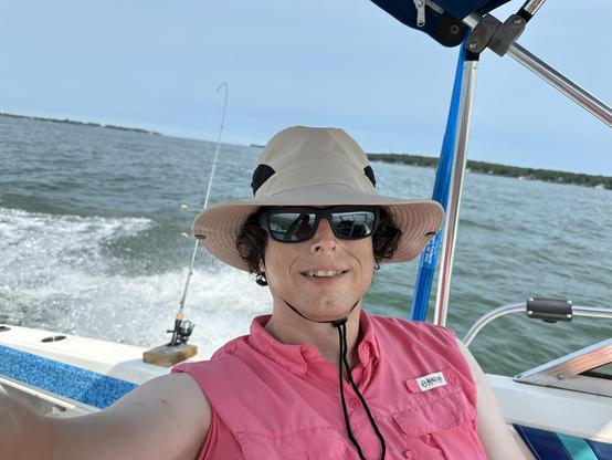 woman in a bright pink outdoor shirt with the sleeves rolled up. she is wearing boring but effective sunglasses and a hat that is large enough for her big head and brain. she is riding in a boat going about 25 mph and you can see the boat's wake in the background