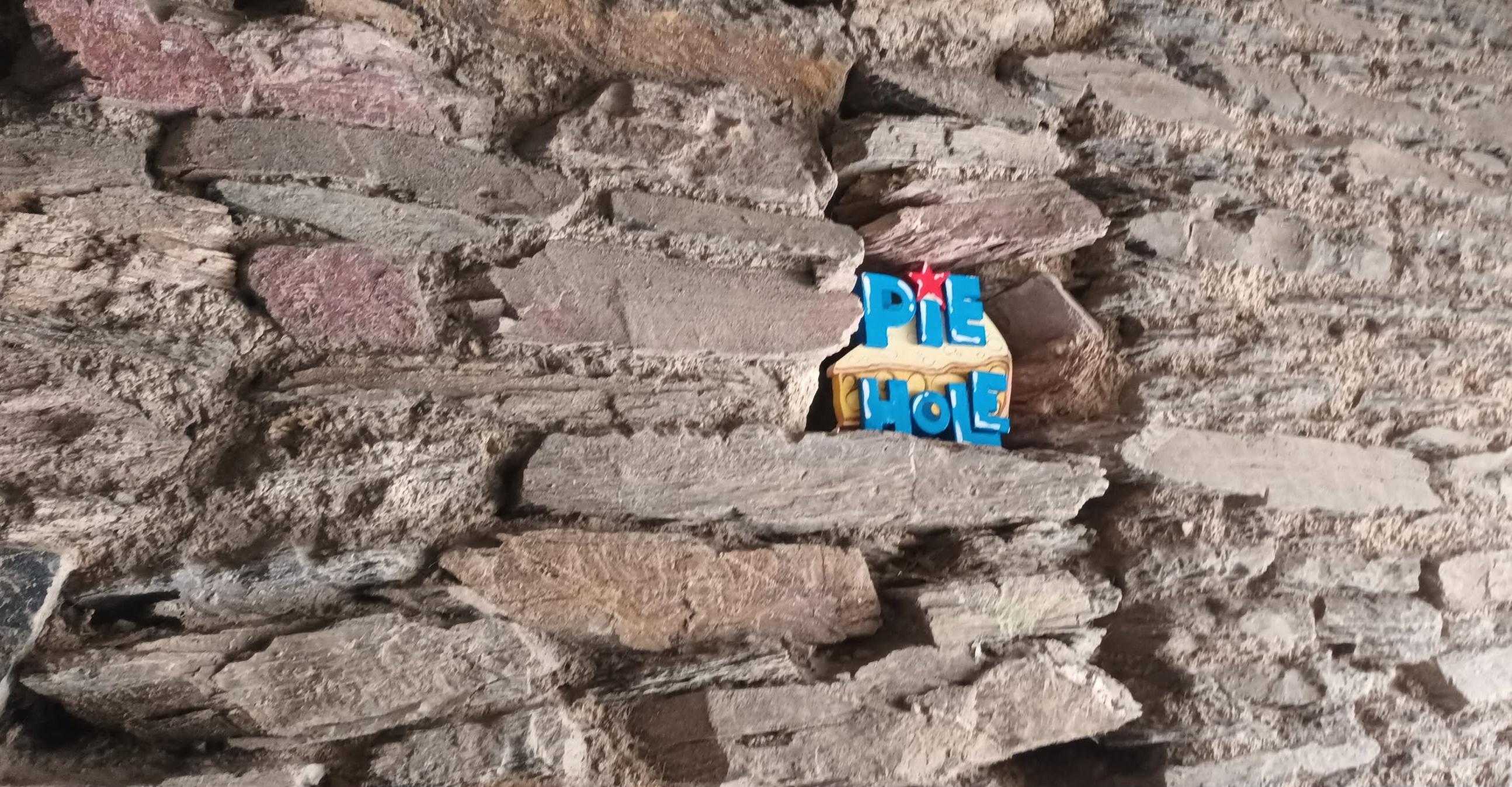 Close up photo of a stone wall in a courtyard. In a small gap between the stones someone has installed a piece of urban art - a 2D triangular slice of cream and brown coloured apple pie. Across the surface of the pie are the words "PIE HOLE" in jaunty blue letters, with a red star instead of a dot above the "i".