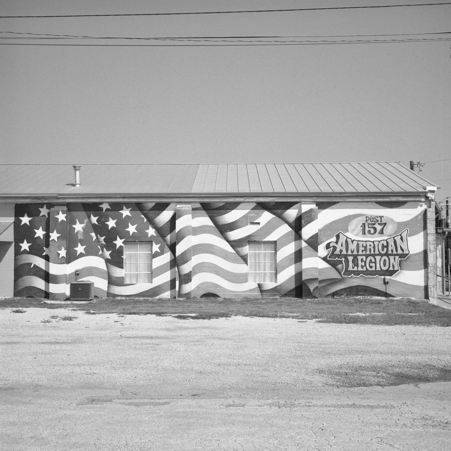 A black-and-white film photo. A giant mural of the American flag can be seen on the side of American Legion Post 157 in Bandera, Texas. Sunday, August 2, 2026.