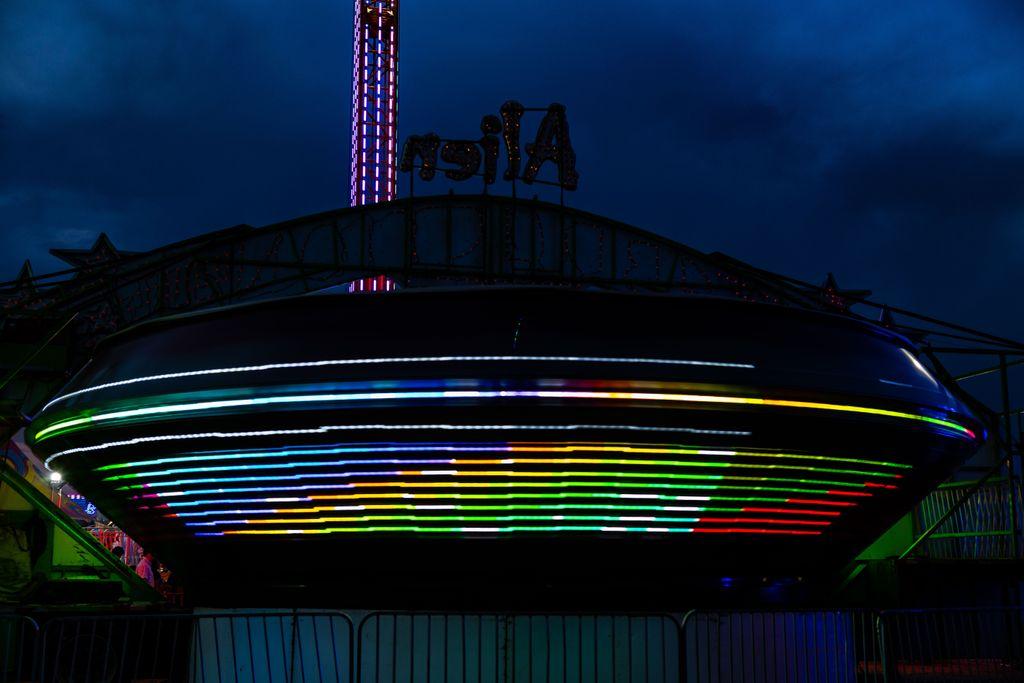 A long-exposure night photo of a spinning saucer-shaped carnival ride at blue hour. The ride’s lights are blurred into horizontal streaks of rainbow color… blue, green, yellow, red, and white… around its dark disc shape. Behind it, a lit drop-tower ride and a sign reading “Alien” (seen in reverse) stand against a deep blue, cloudy sky. A fence runs along the bottom of the frame.