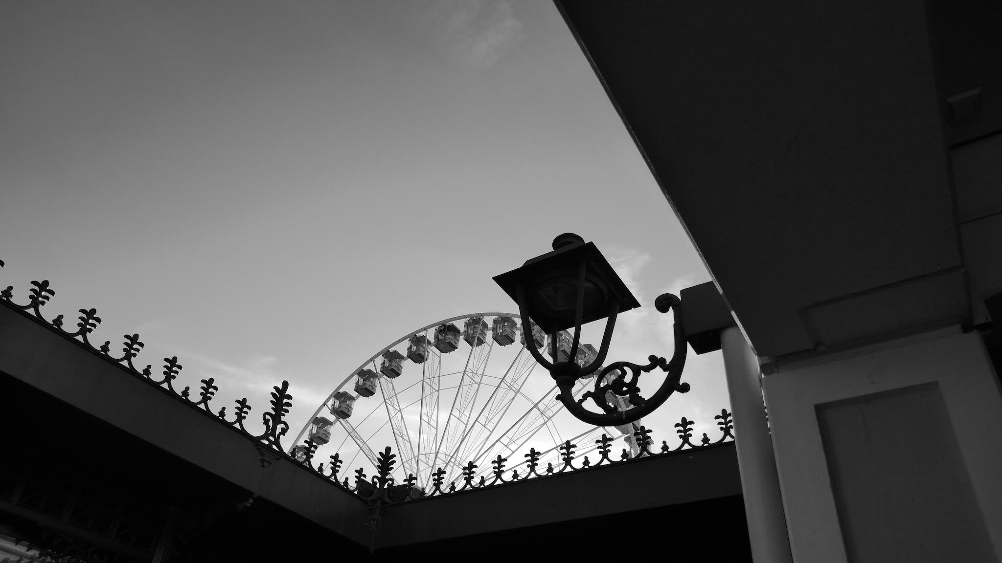 Picture shows a part of giant wheel in the background. In the center of the photo is shape of lantern, mounted on a roof. A floral pattern of small metallic ornaments are the button frame. Picture is black and white.