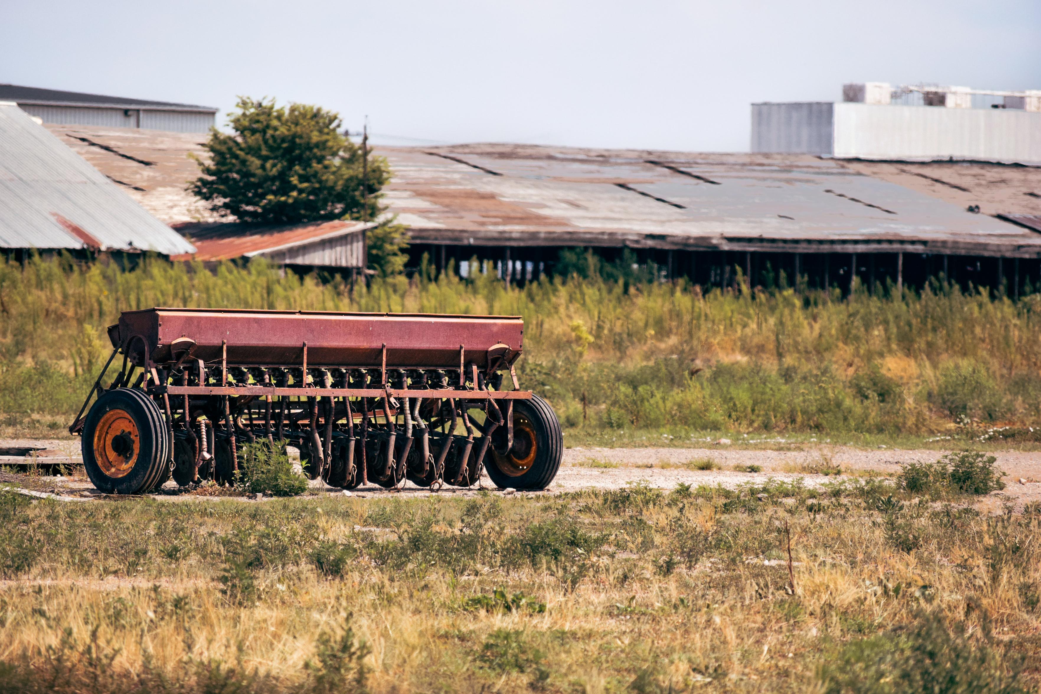 An old red farm machine with large black wheels sits on dry, grassy land. Behind it, rusty warehouses and greenery create a neglected, rustic atmosphere.