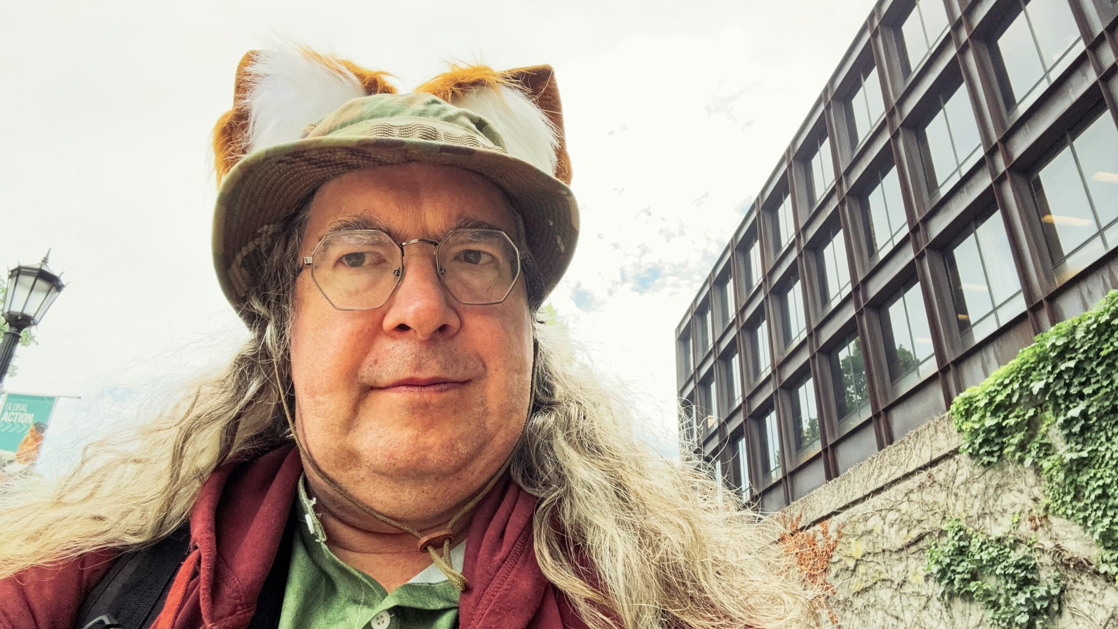 Middle aged man with a lot of curly hair flowing around his shoulders is wearing a green shirt and maroon hoodie and a boonie cap with two orange and white fox ears on top;  to the left you can see a bit of a street lamp like the one the witch trashed in Narnia and on the right a building that wants to be seen in two point perspective.