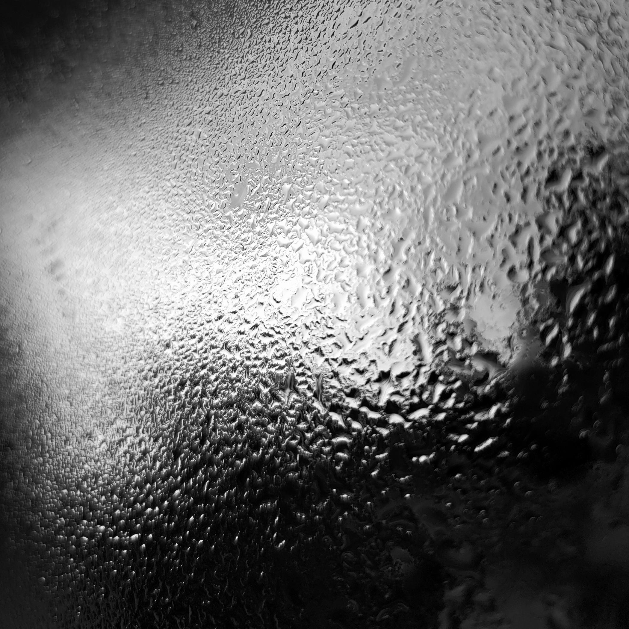 B&W close up of rain speckled glass, with bright diffused light glowing behind it and deep shadows creating an abstract texture.