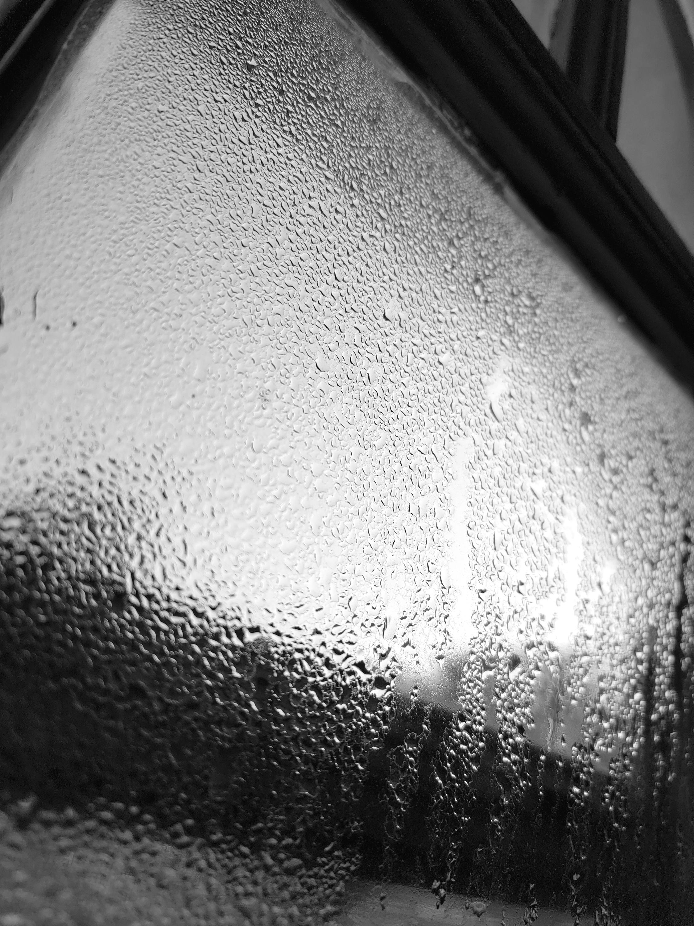 Monochromatic photo of condensation and raindrops covering a window, with bright light and dark window framing.
