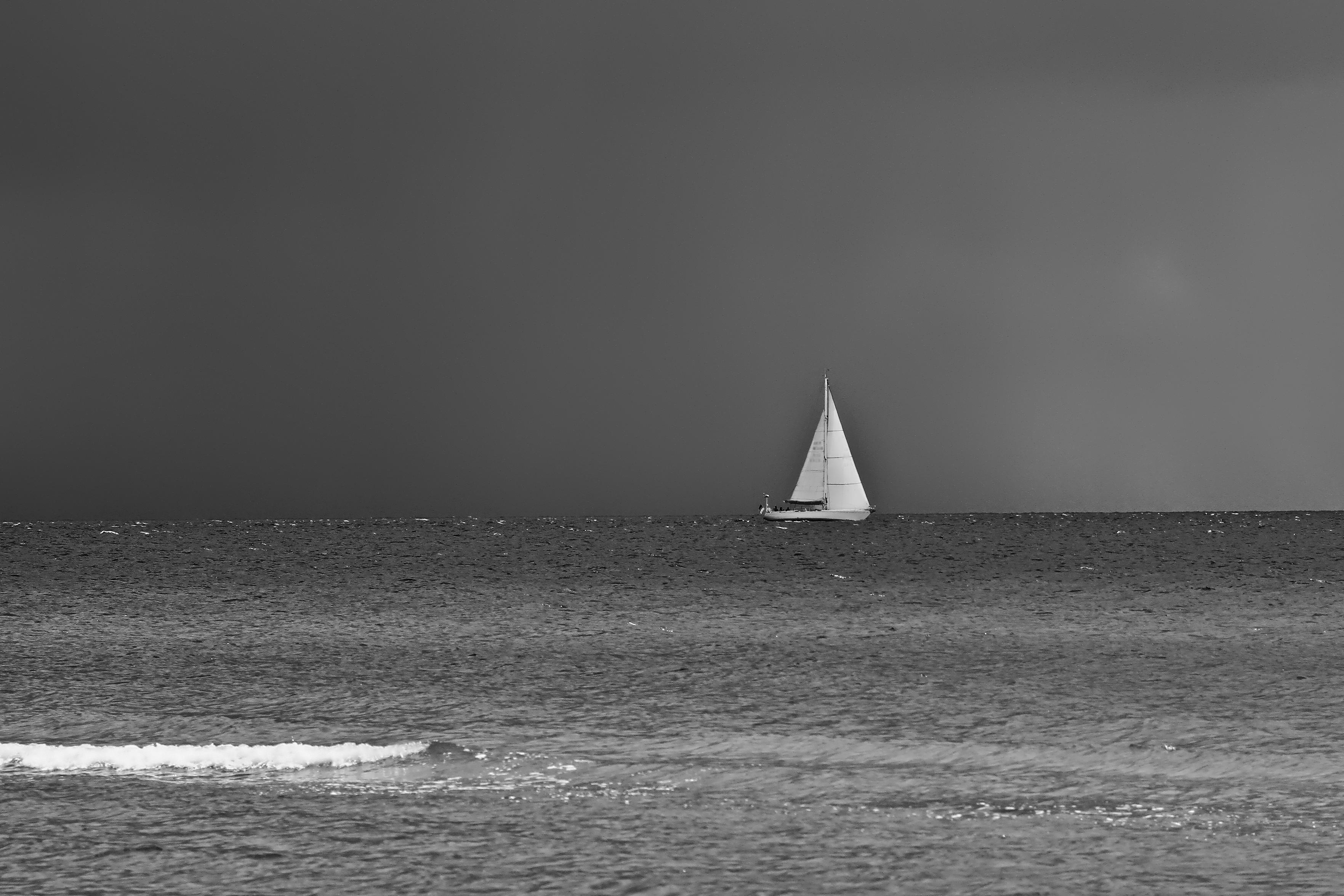 Schwarzweiss-Foto der küstennahen Ostsee mit dunklen Wolken am Himmel. In der Mitte ungfähr auf Höhe des Horizontes ist ein Segelboot zusehen, das noch von der Sonne angeschienen wird.