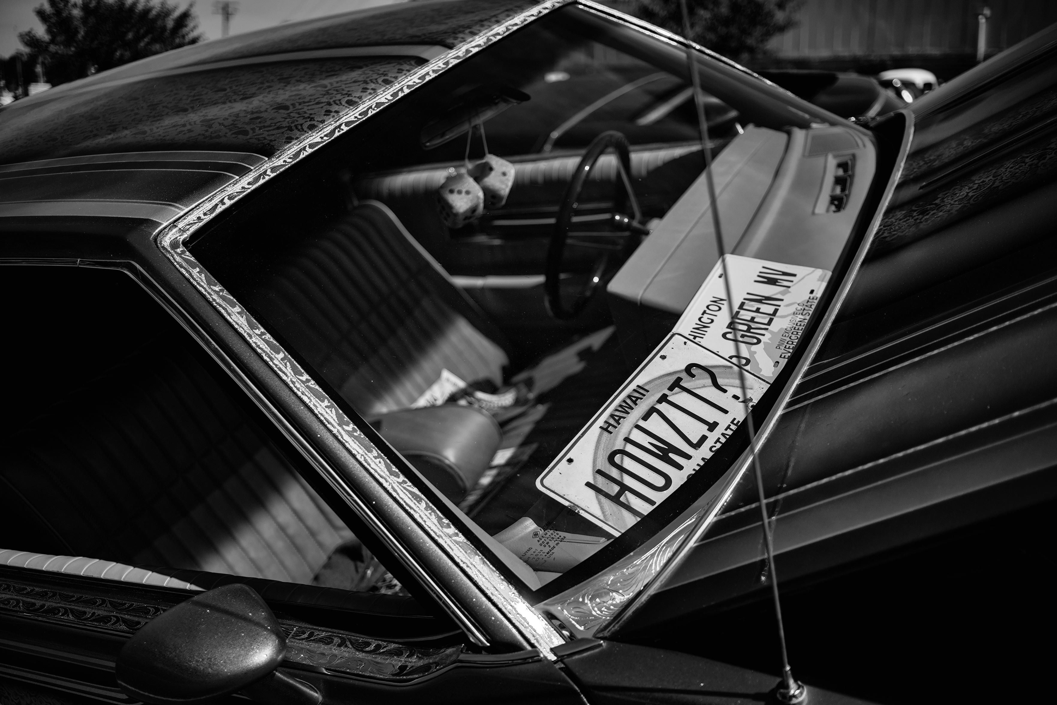 Black and white photo of the dashboard in landscape format of two vanity license plates prominently displayed on top the inside dashboard of a custom car through the front windshield.

The Hawaii license plate is on the passenger side with the word "HOWZIT?" in all caps vertically and horizontally centered and taking up two thirds of the plate. Above that, vertically and horizontally centered in all caps is the word "HAWAII" with font about a quarter size compared to "HOWZIT?". A rainbow arches from the bottom left and right edges of the plate. The bottom of the plate has a barely visible "ALOHA STATE" between the bottom edges of the rainbow.

The left third of the other license plate is underneath the Hawaii plate and not visible. What is visible: "HINGTON" on one "line, "GREEN MV" next, "PNW EXCLUSIVE C.C." next and " EVERGREEN STATE" on the bottom.

The rest of the interior is slight out of focus due to depth of field. A dark colored couch seat with middle divider, dark colored stee