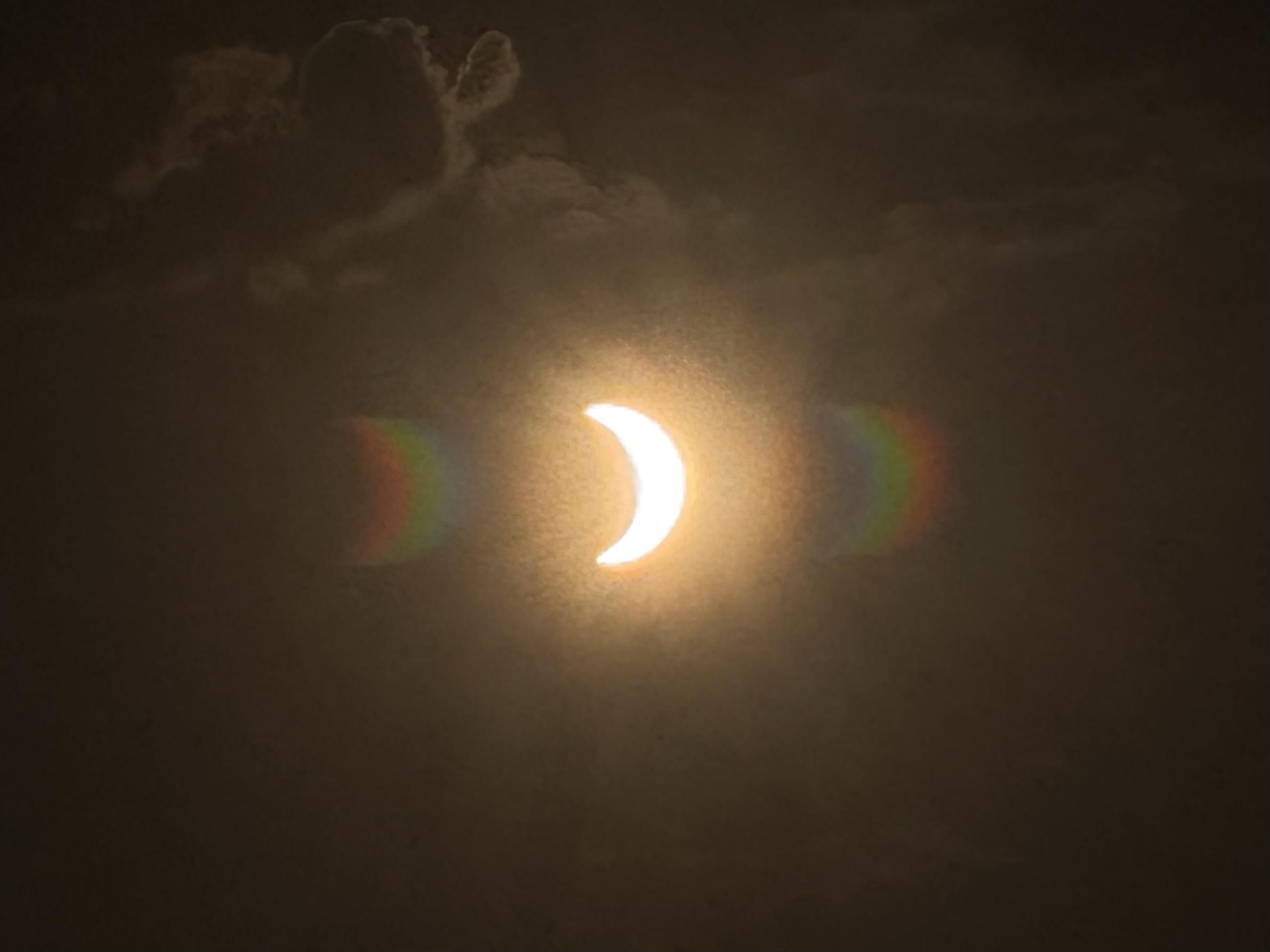 A partially eclipsed sun against a cloudy sky, with colorful lens flare on either side the same shape as the sun.