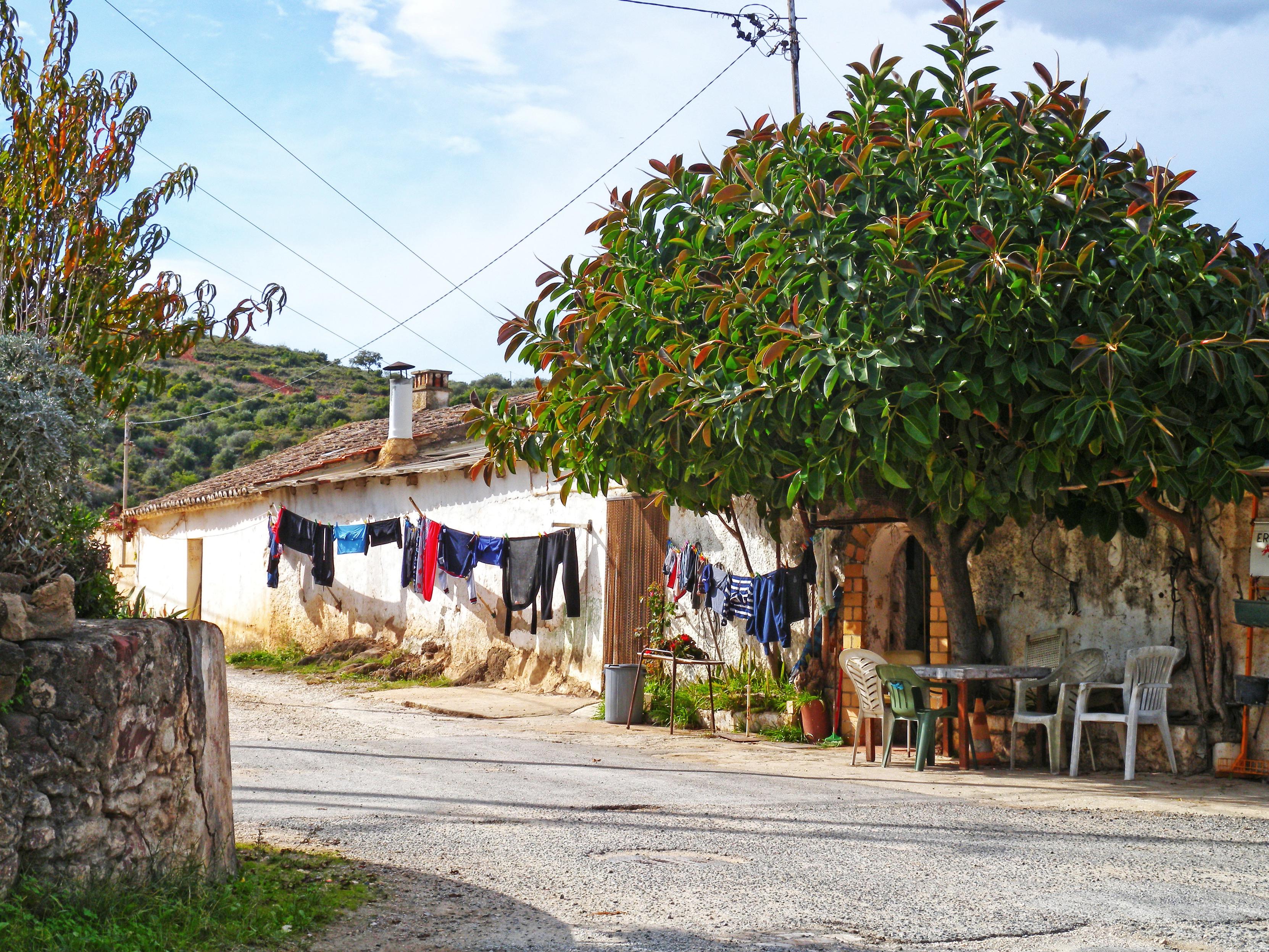 A cena mostra uma casa rural caiada de branco, com telhado de telhas e varal à beira da estrada, numa região montanhosa ensolarada. À direita, uma árvore grande e frondosa dá sombra a um pequeno conjunto de mesas e cadeiras ao ar livre. Ao fundo, estende-se uma paisagem verdejante e ondulada sob um céu azul.
