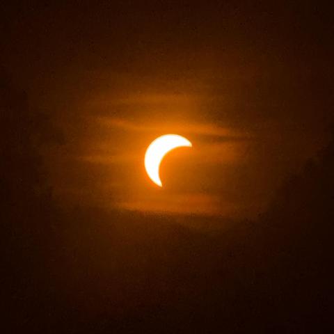 Bild von der Sonne durch die Schutzbrille, Sonnenscheibe in orange, der Mond bedeckt 50% bei der partiellen Sonnenfinsternis. Sieht aus wie eine Sichel. Drumherum schön orange-braun eingefärbte Schleier in der Luft.